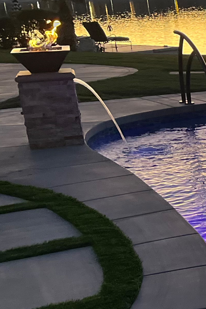 A small waterfall seamlessly integrated into a swimming pool, featuring a fire element above it, all set against a scenic lake view.