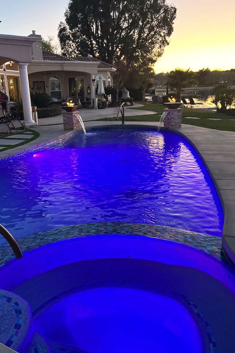 A beautiful blue swimming pool with an integrated jacuzzi, offering stunning views of a lake and an elegant house.