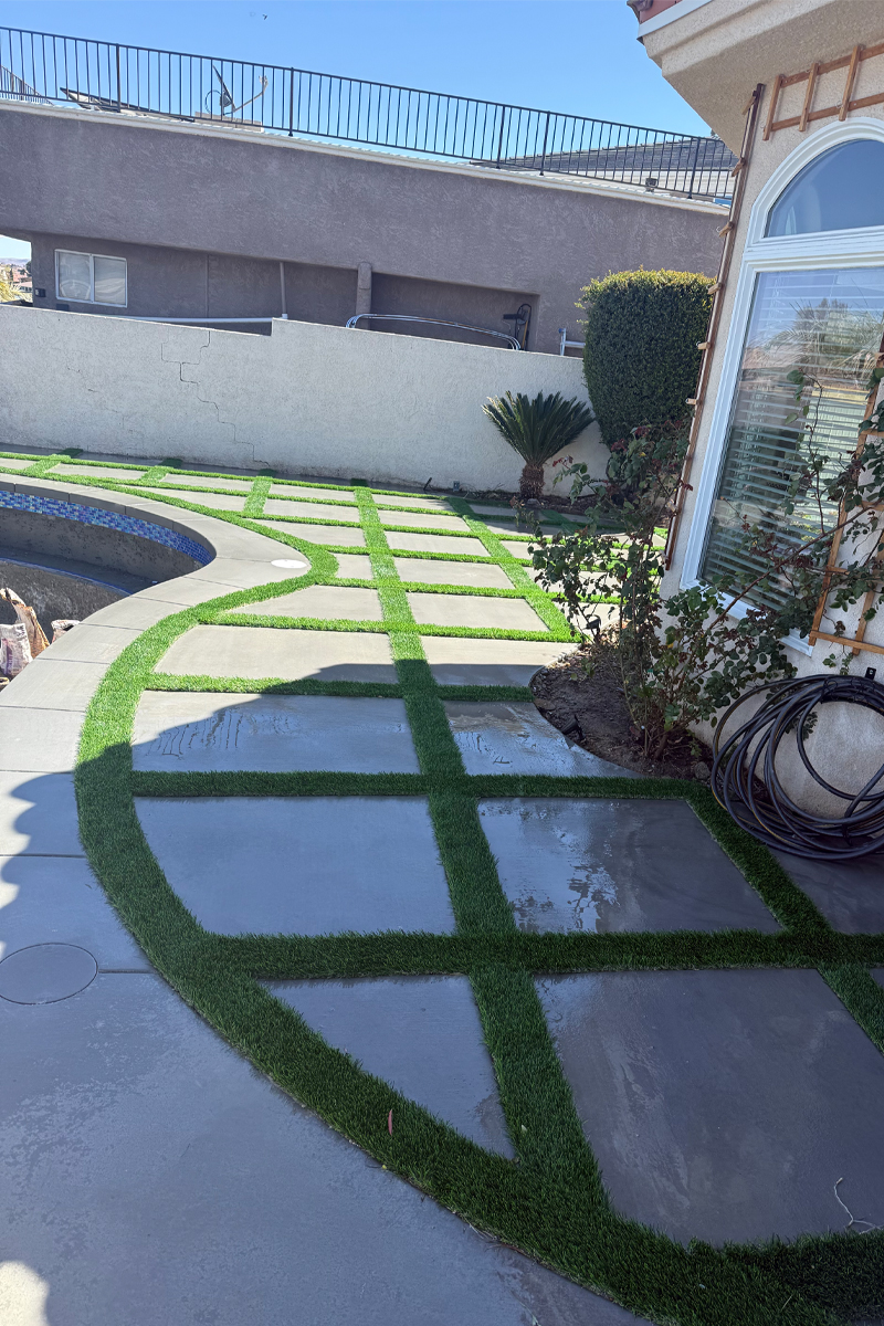 A newly finished patio composed of square sections of fresh cement, seamlessly integrated with lines of artificial grass.
