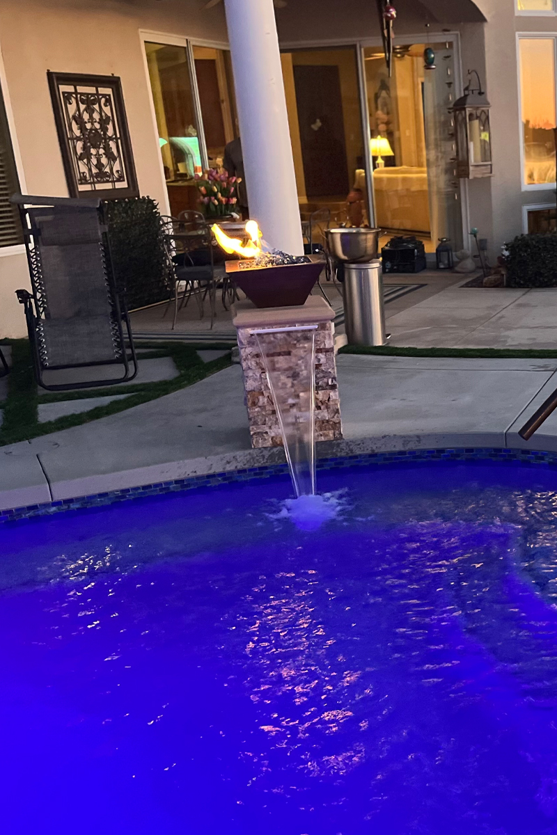 An intense blue swimming pool featuring a waterfall and a base with fire burning on top. The background shows a view of the house's living room, suggesting an integrated indoor-outdoor design.