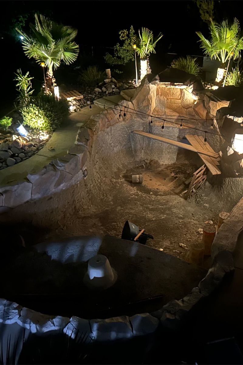 The framework of a swimming pool is under construction, surrounded by decorative stones and several palm trees.