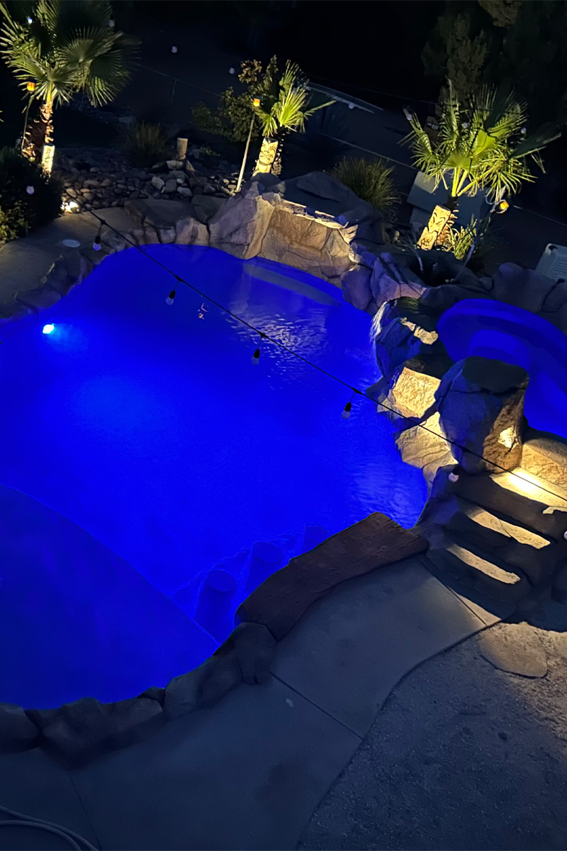 A beautiful, brightly illuminated swimming pool with intense blue water. The pool is made of stone and surrounded by palm trees.