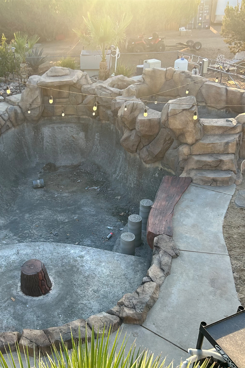 The framework of a swimming pool is under construction, surrounded by decorative stones and several palm trees.