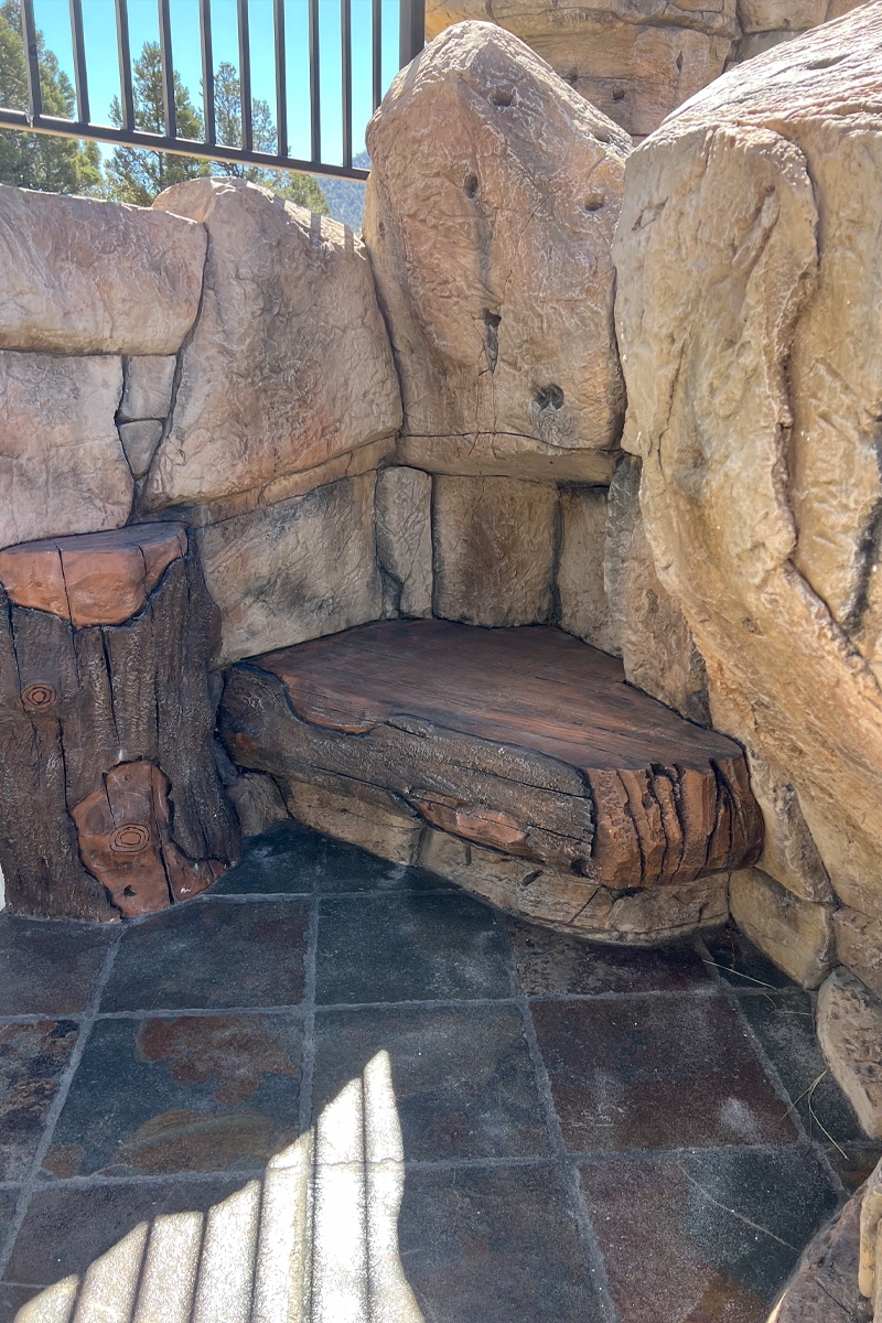 A unique seating area featuring a rustic seat made from a tree trunk or large branch, which is firmly embedded into a base of natural stones.