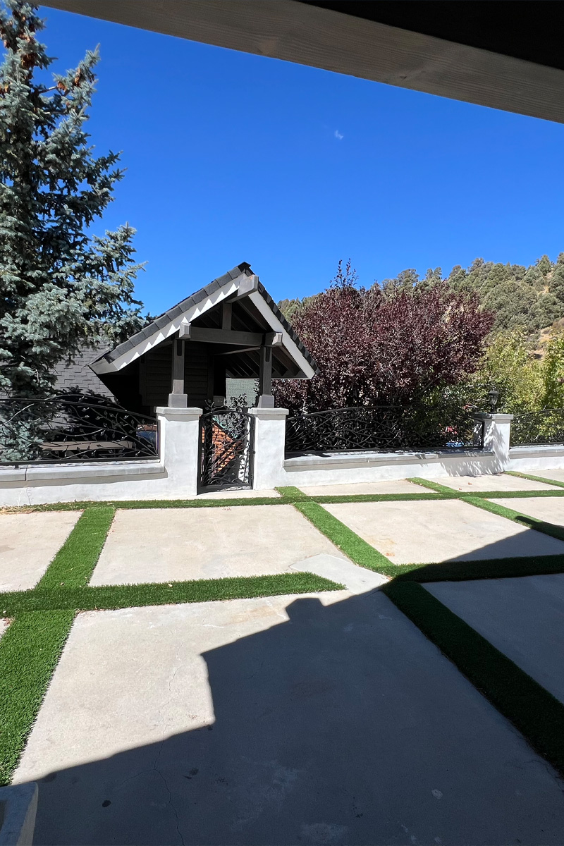 A patio made of cement, featuring integrated lines of artificial grass. From this patio, there is a clear view of an entrance door crafted from metal with decorative designs.