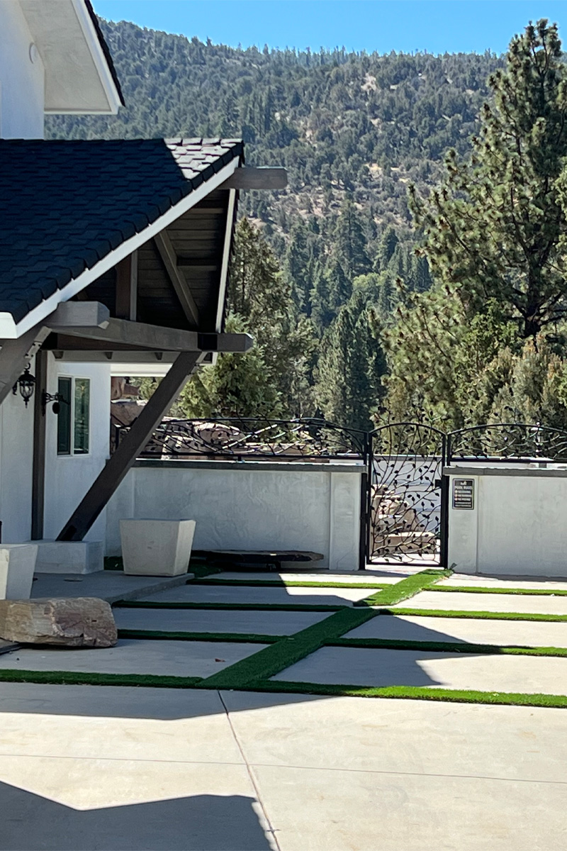 A view from the interior looking out onto a modern patio. The patio floor is made of cement with integrated lines of artificial grass, creating a clean, geometric pattern. In the background, there's a beautiful vista of pine trees on a mountain.