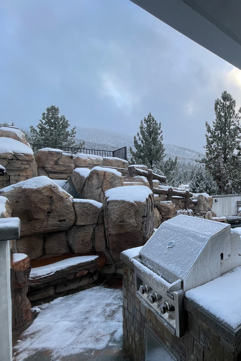 A beautiful patio featuring a fully equipped outdoor kitchen. The background showcases a scenic view of natural stone formations and pine trees covered in snow, creating a charming winter outdoor living space.