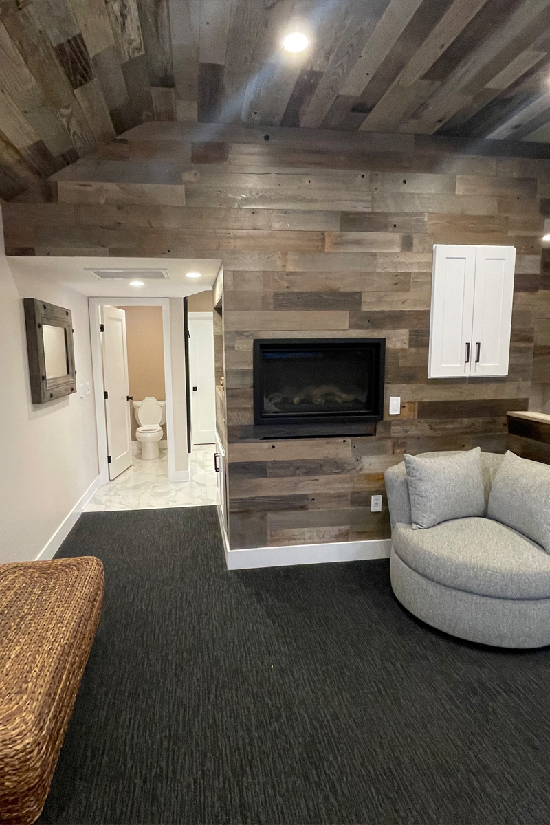 A beautiful living area seamlessly integrated into a bedroom. The ceiling and walls are made of warm wood paneling. A comfortable grey sofa is placed in the living space, and a glimpse of a bathroom can be seen in the background.

