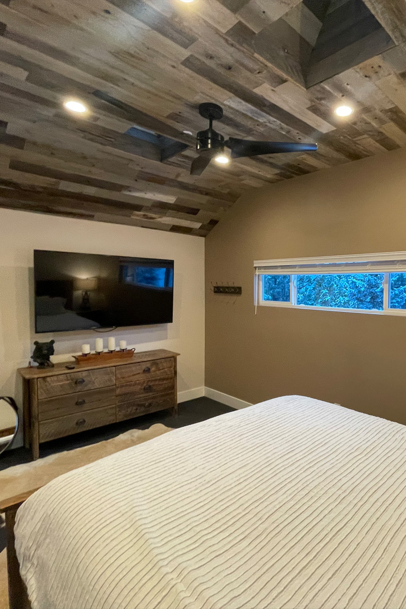 A beautiful bedroom featuring a stylish grey wooden ceiling. A television is mounted on the wall, and a tall, slender window allows natural light into the room. A sophisticated wooden furniture piece adds an element of elegance and refinement.