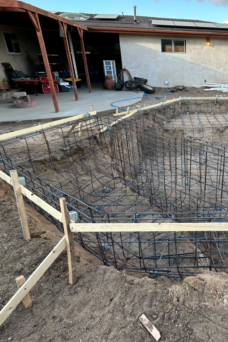 An in-ground swimming pool under construction. The interior is a concrete shell, and the surrounding deck area is framed with a mixture of wood and metal.
