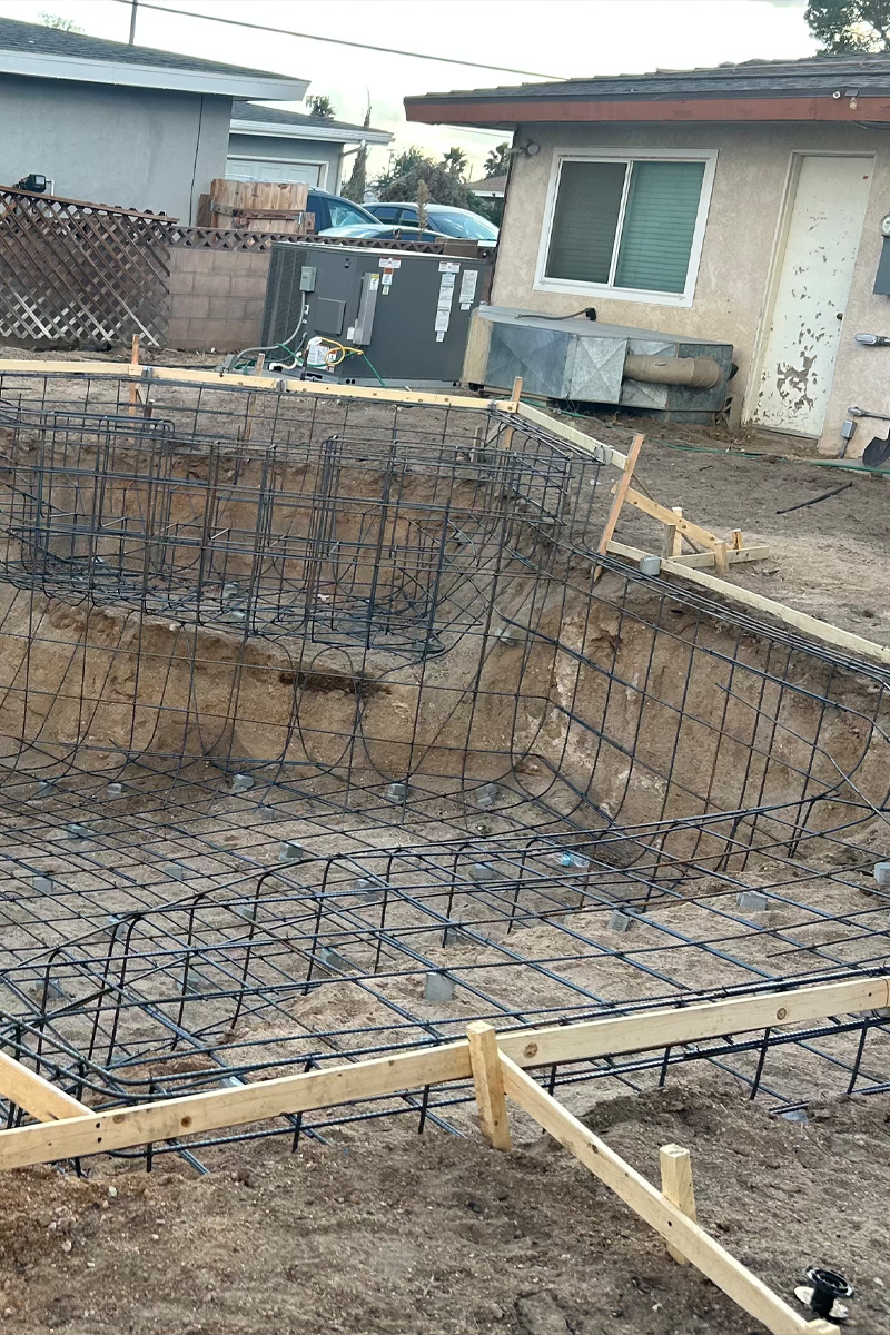 An in-ground swimming pool under construction. The interior is a concrete shell, and the surrounding deck area is framed with a mixture of wood and metal.