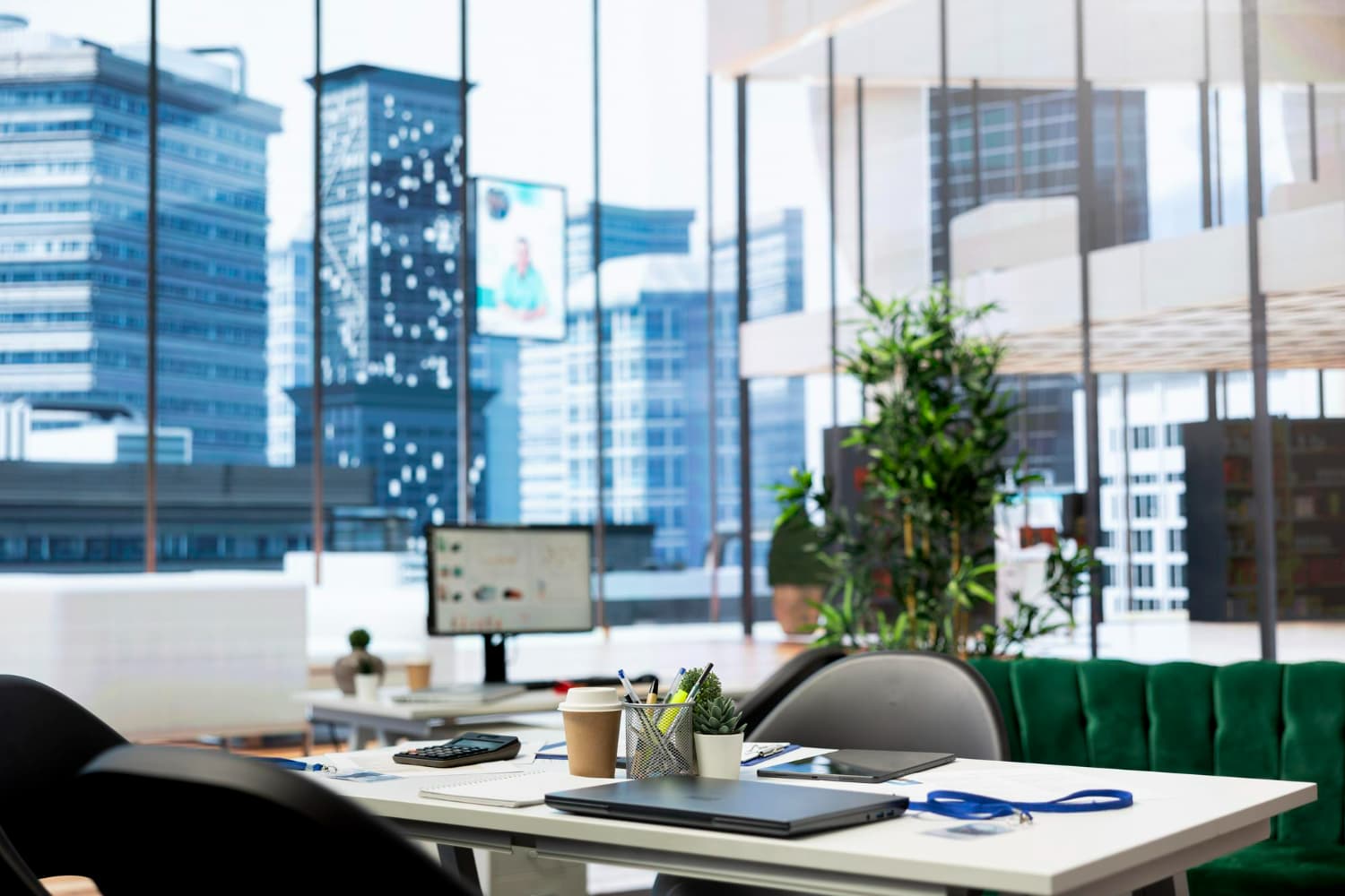 Modern office desk with laptop, coffee cup, pens, and plants near large windows showing city buildings.