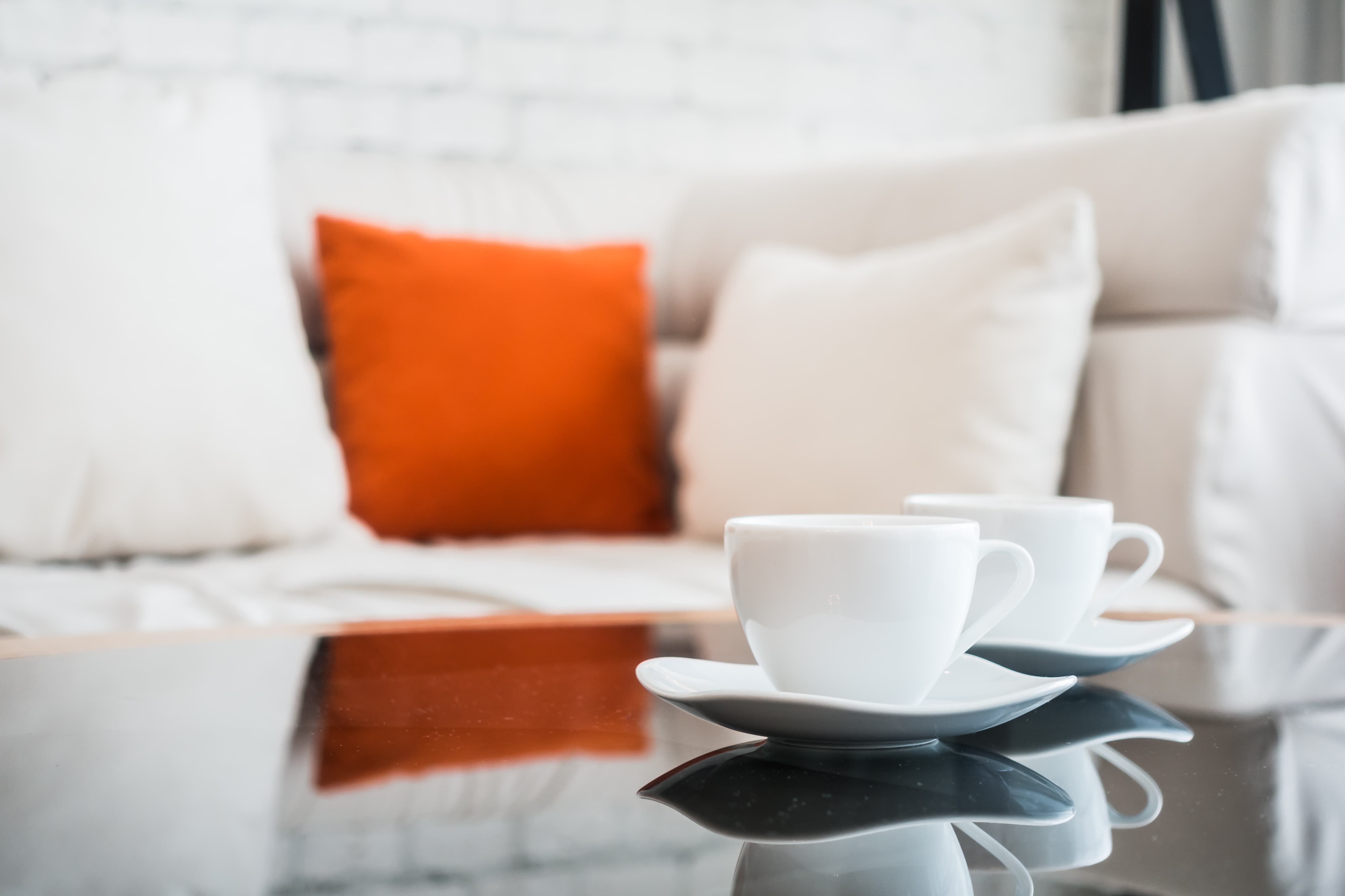 Two white coffee cups with saucers on a glass table in front of a white couch with colorful pillows.