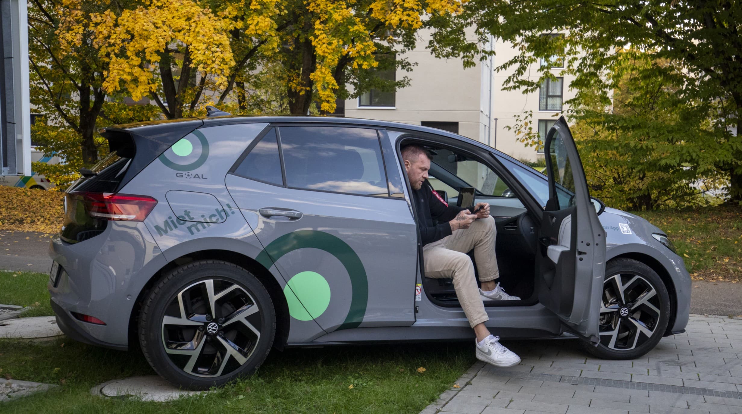Man sitting in driver's seat of a grey Volkswagen car with open door, checking his phone.
