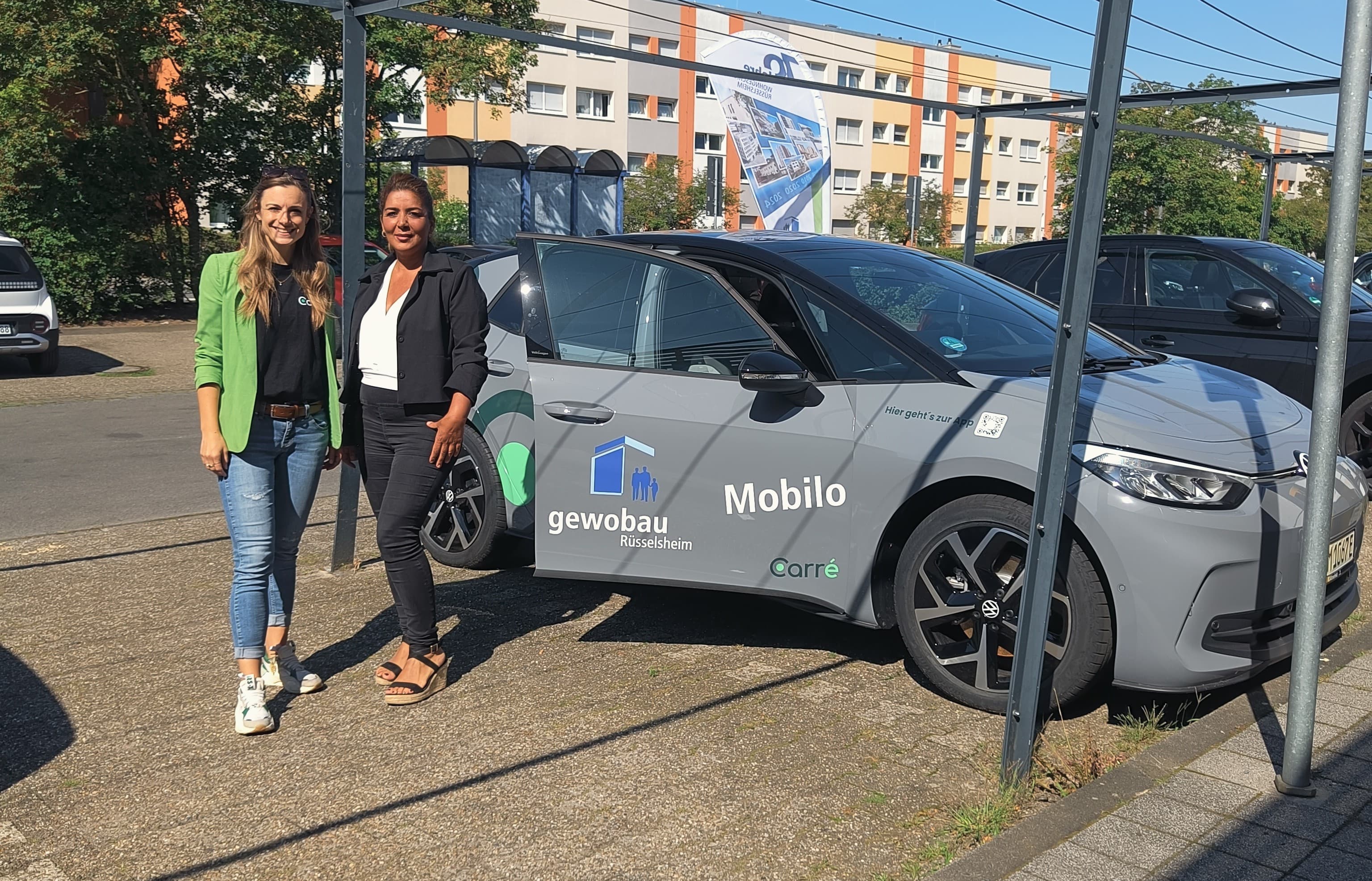 Two women standing next to a gray electric car with gewobau Rüsselsheim and Mobilo logos.
