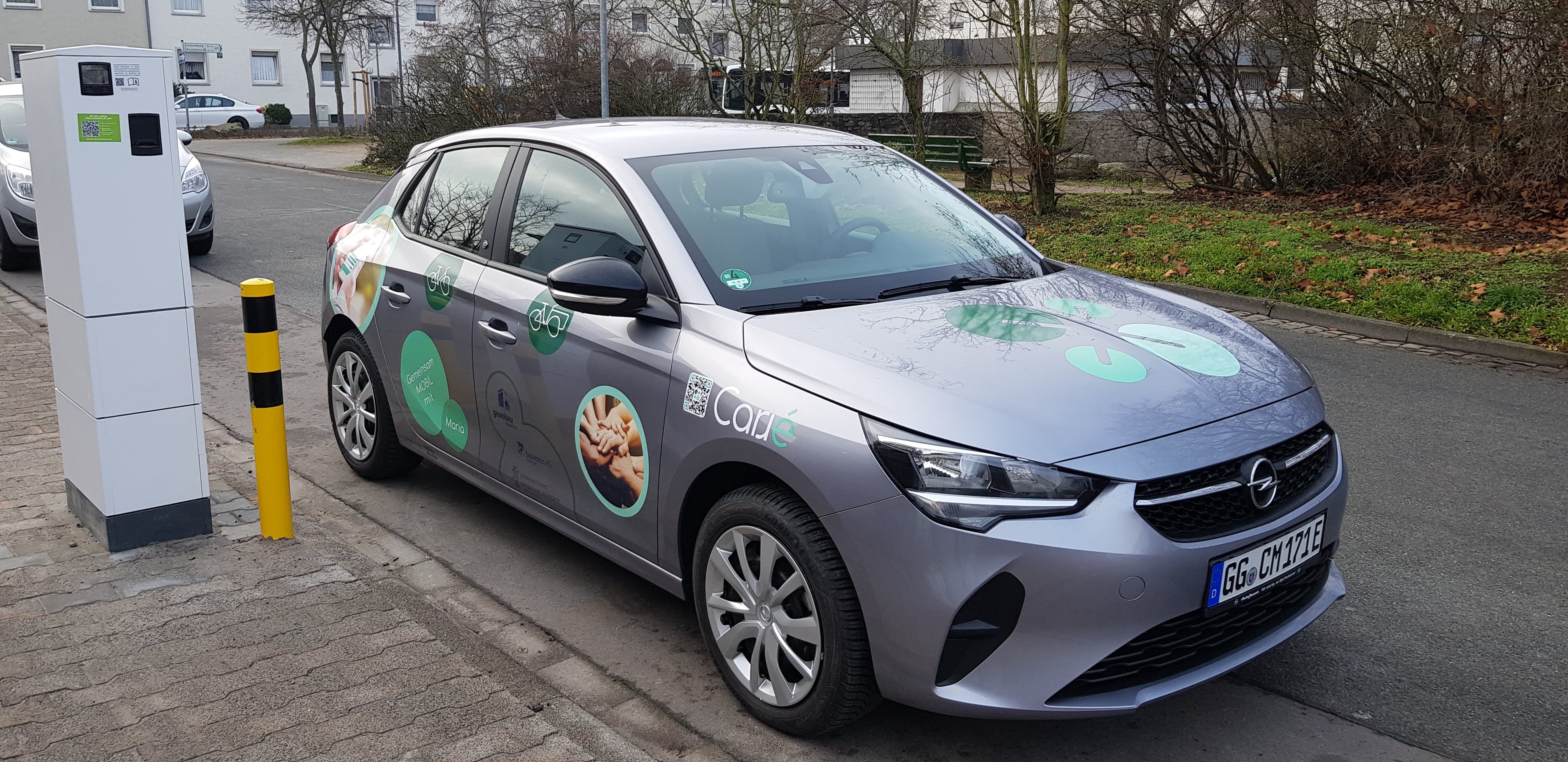 Silver Opel car with green circular decals parked by a curb near a white charging station.