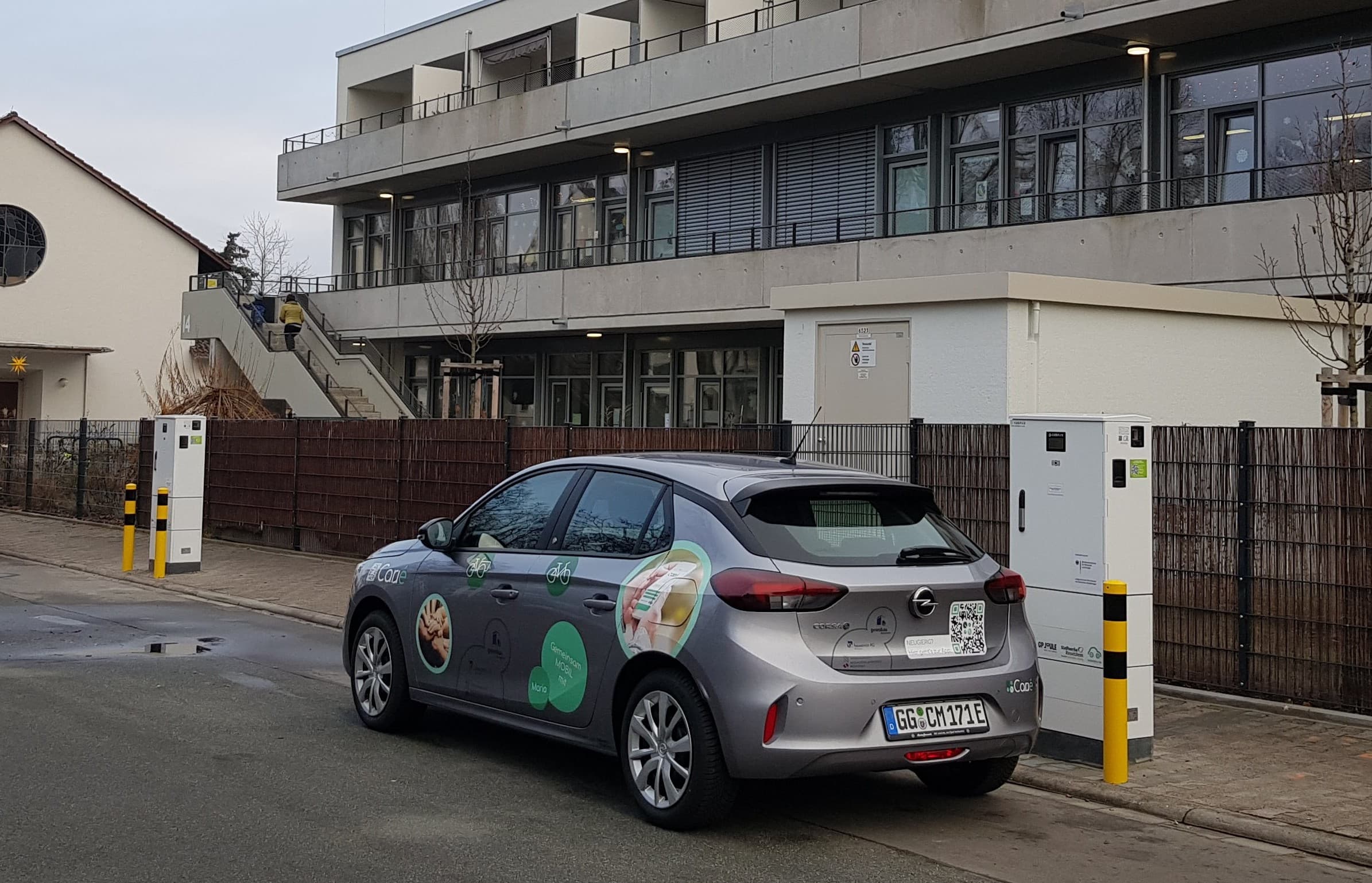 Gray Opel hatchback with promotional decals parked next to a charging station on a city street.
