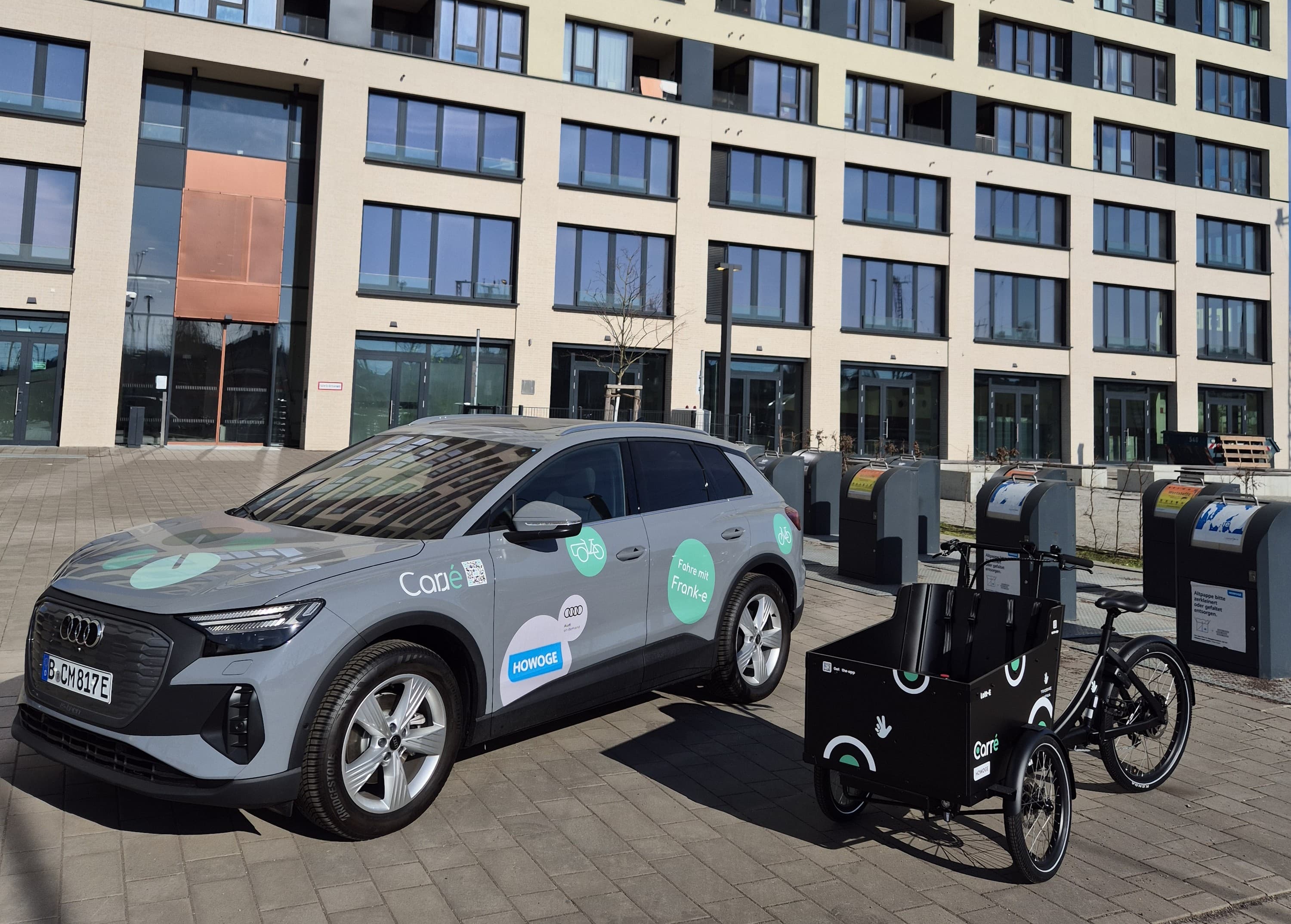 Gray Audi SUV with logos parked next to a black cargo tricycle outside a modern building.