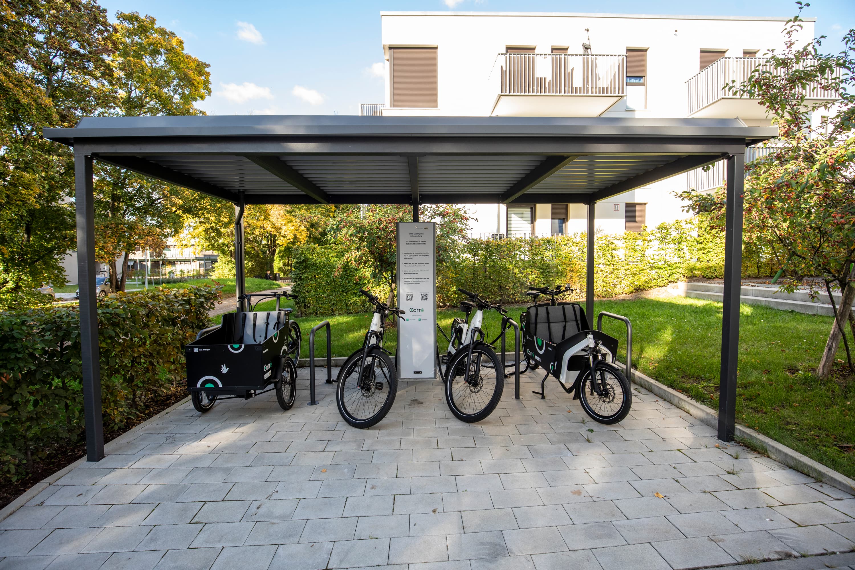 Three electric cargo bikes parked under a metal shelter near a modern building in a green area.