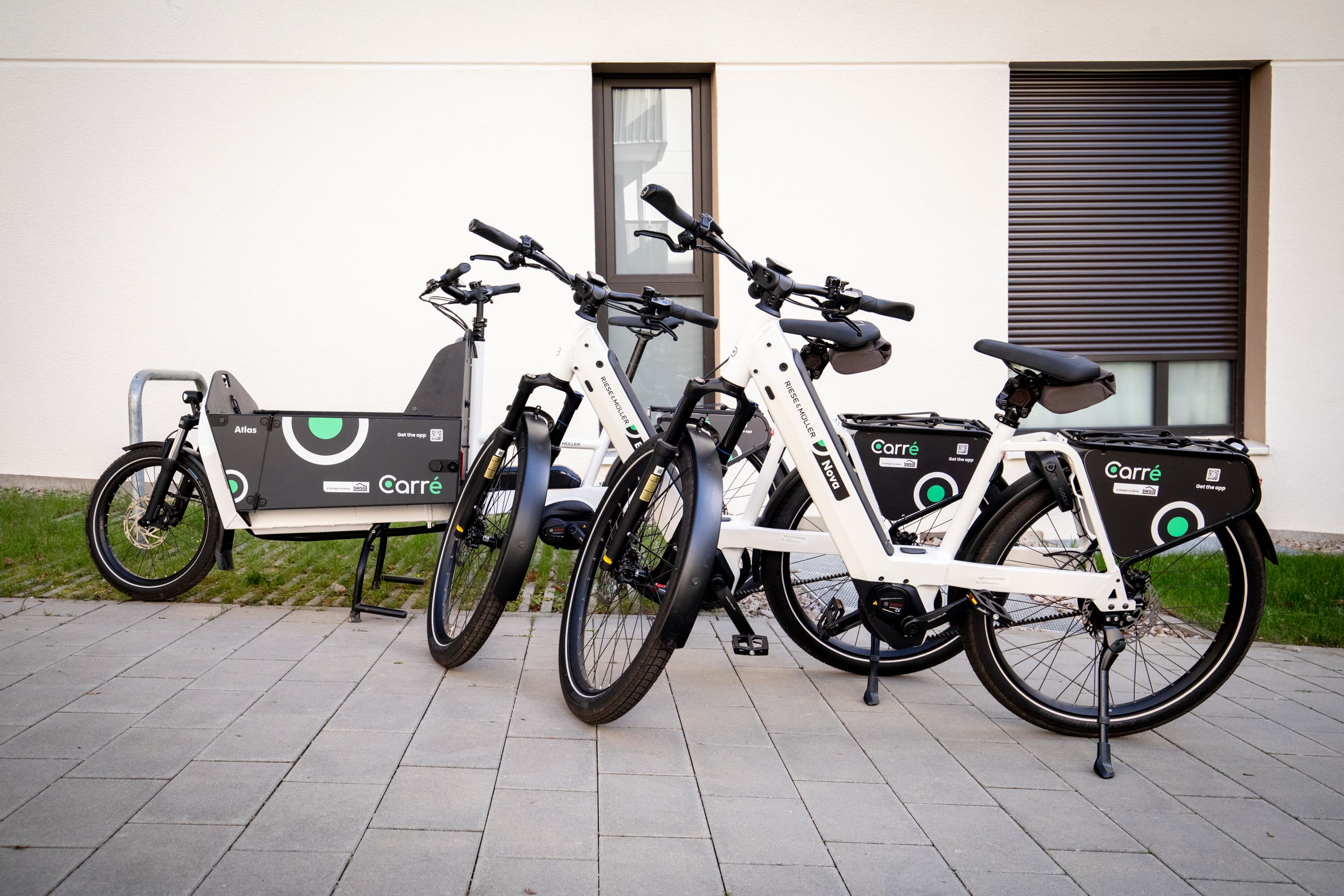Three white Carre electric bikes parked on gray pavement against a white building with windows.