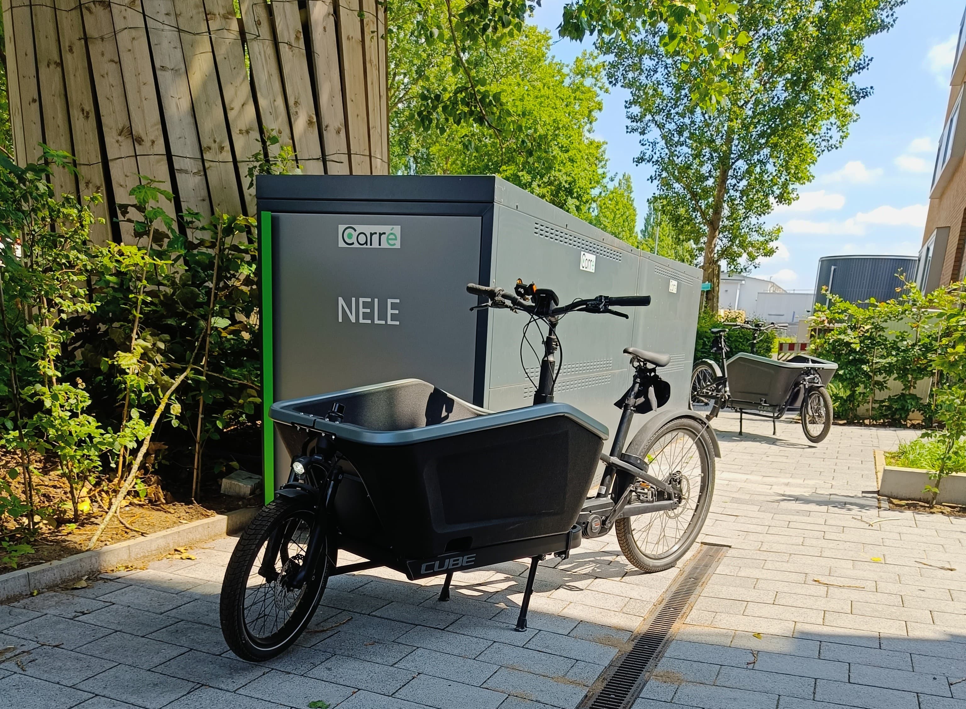 Two black cargo bikes parked on a paved path beside a large box labeled NELE and Carré.