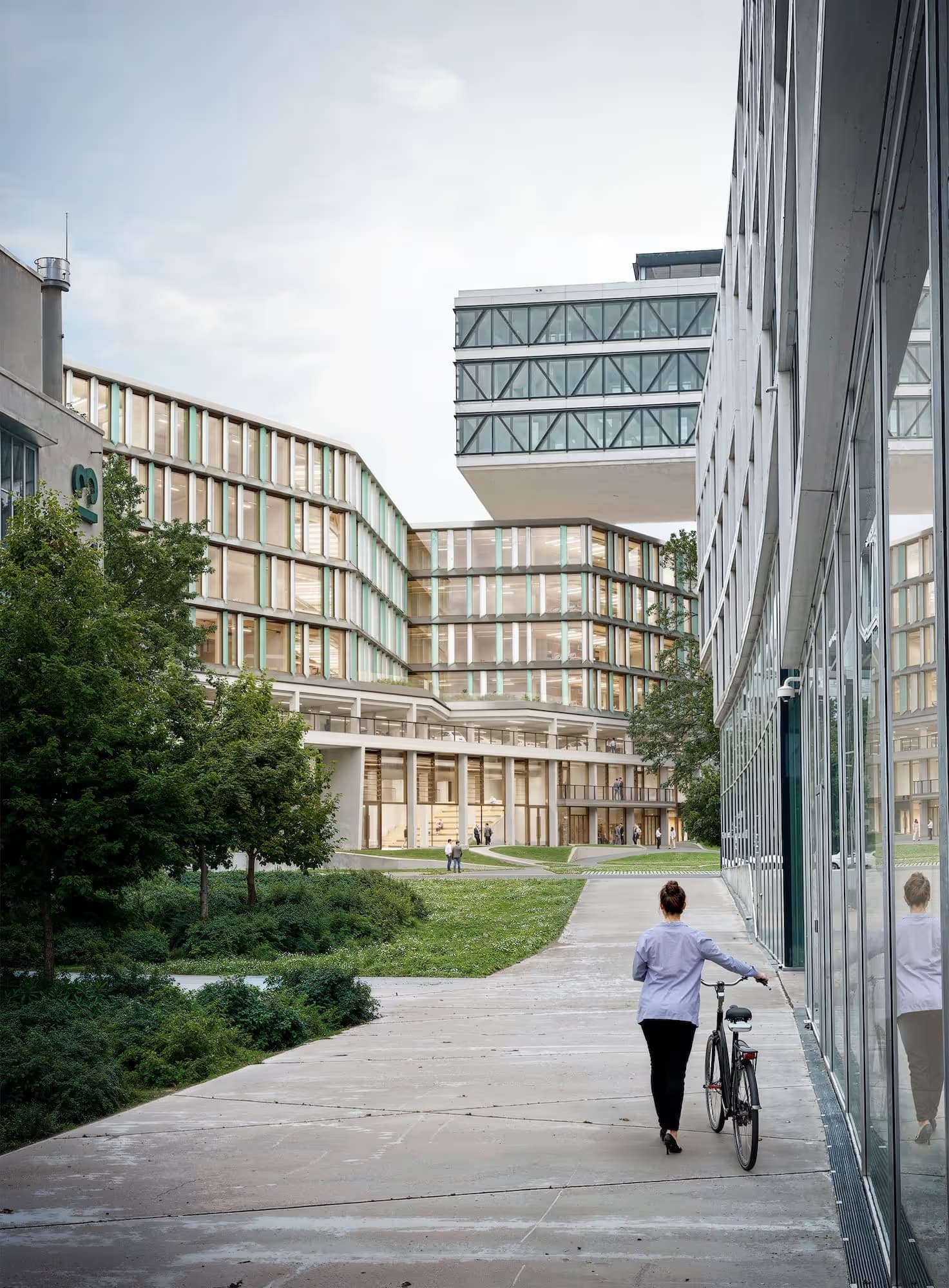 Modern office building with glass windows surrounded by trees, grass, and people walking and biking.