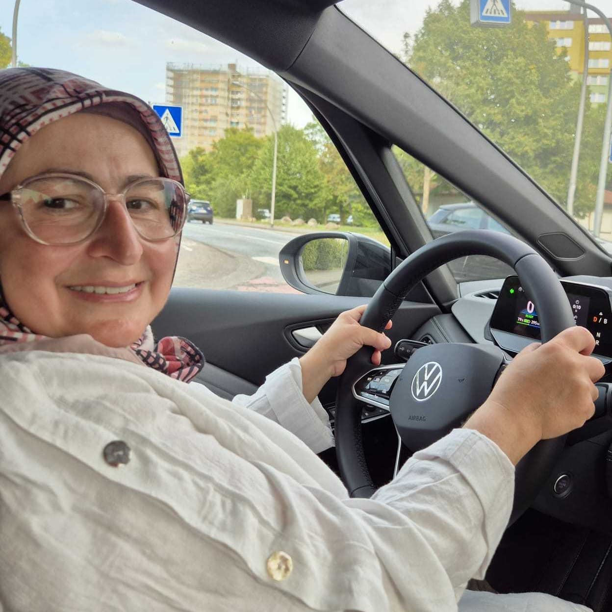 Woman wearing glasses and a headscarf smiling while driving a Volkswagen car on a city street.