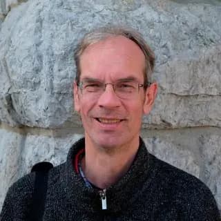 Smiling middle-aged man with glasses and a dark sweater standing outdoors by a stone wall.