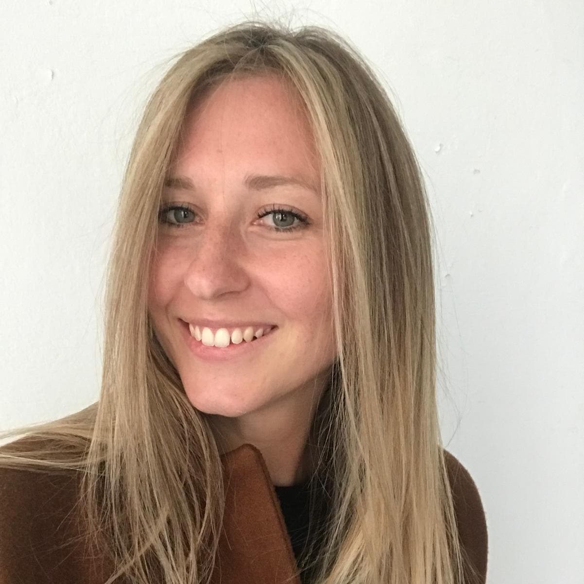 Smiling young woman with long blonde hair wearing a brown coat against a white wall.