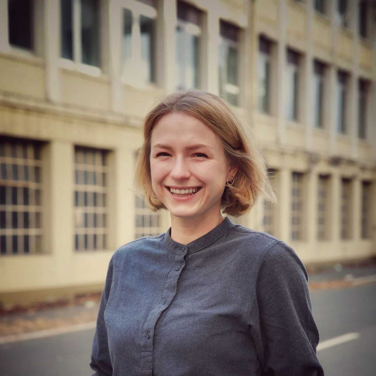 Smiling woman with short blonde hair wearing a dark buttoned shirt standing outdoors near a building.