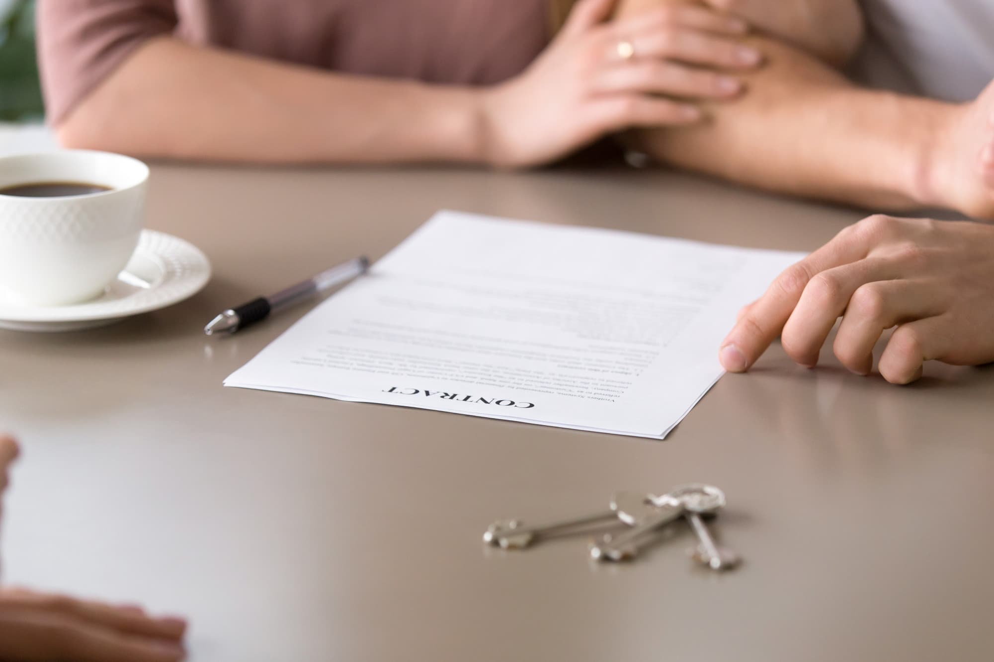 Person pointing to a contract document on a table with keys, a pen, and a coffee cup.