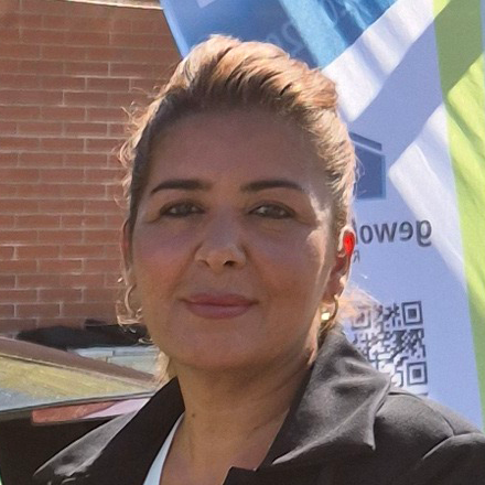 Woman with light brown hair pulled back wearing a black jacket, standing outdoors by a brick wall and banner.