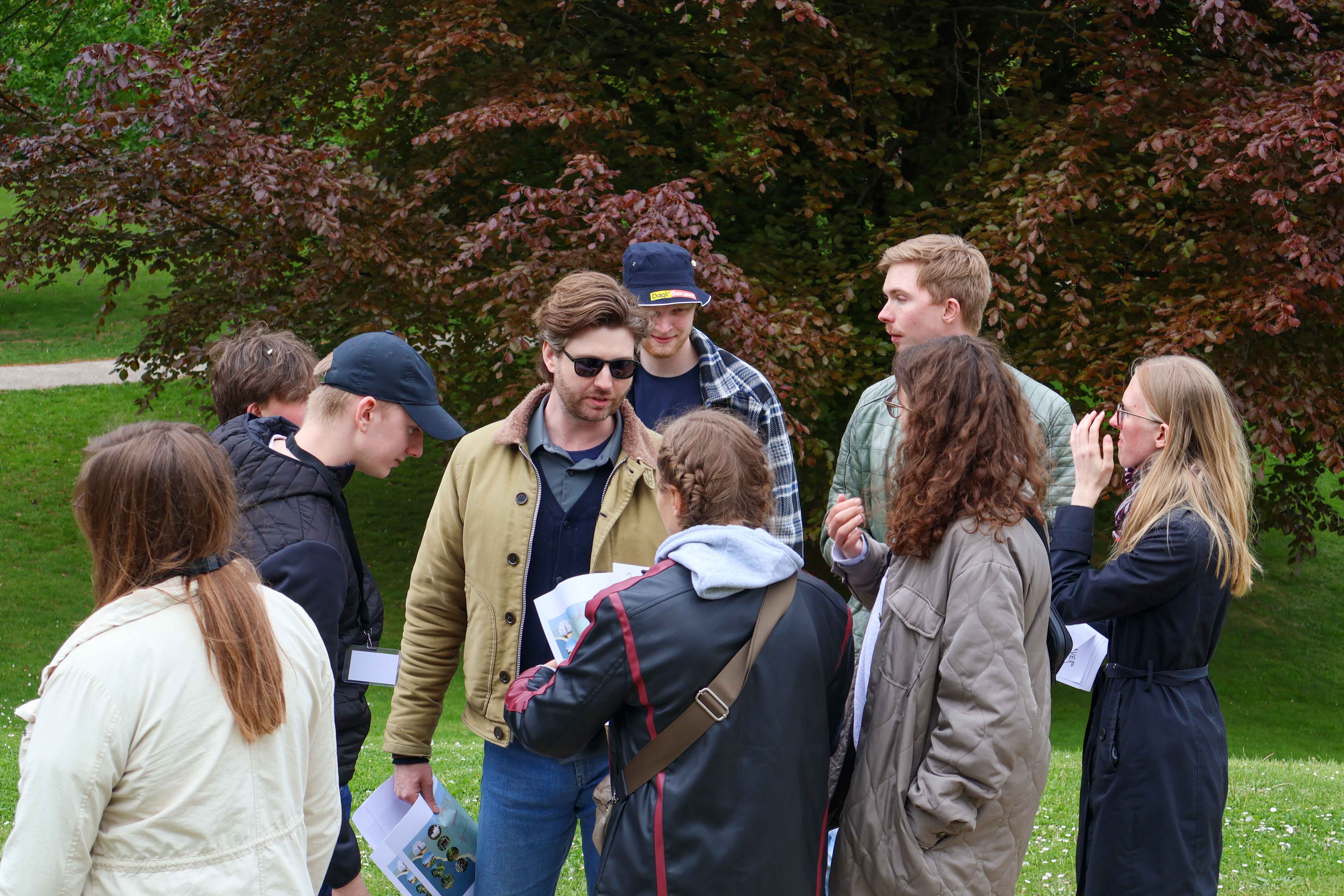 To unge sidder sammen forrest i en rød rutsjebanevogn formet som en bille med øjne og små dannebrogsflag. De ser glade og spændte ud, omgivet af farverige blomster og træer på en solrig dag i en forlystelsespark.