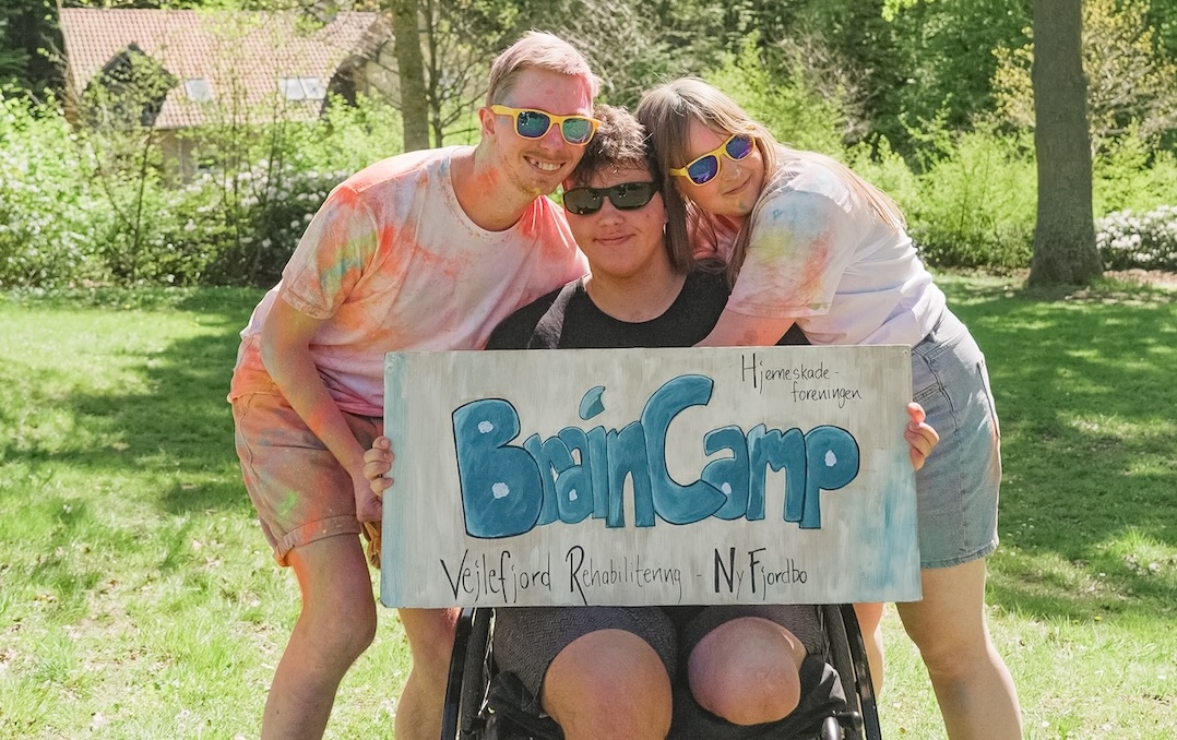 Et gruppebillede af omkring 35 unge voksne, som smiler og hygger sig udendørs på en græsplæne under et træ, med et skilt foran hvor der står ’BrainCamp’.