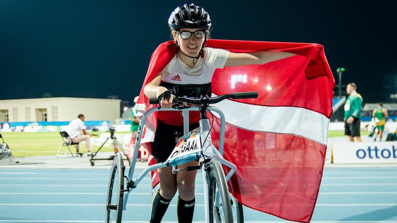 Billede af en ung atlet står med et stort smil på løbebanen iført hjelm og specialcykel. Personen er indhyllet i et dannebrogsflag og omgivet af sportsstemning under stadionlys.