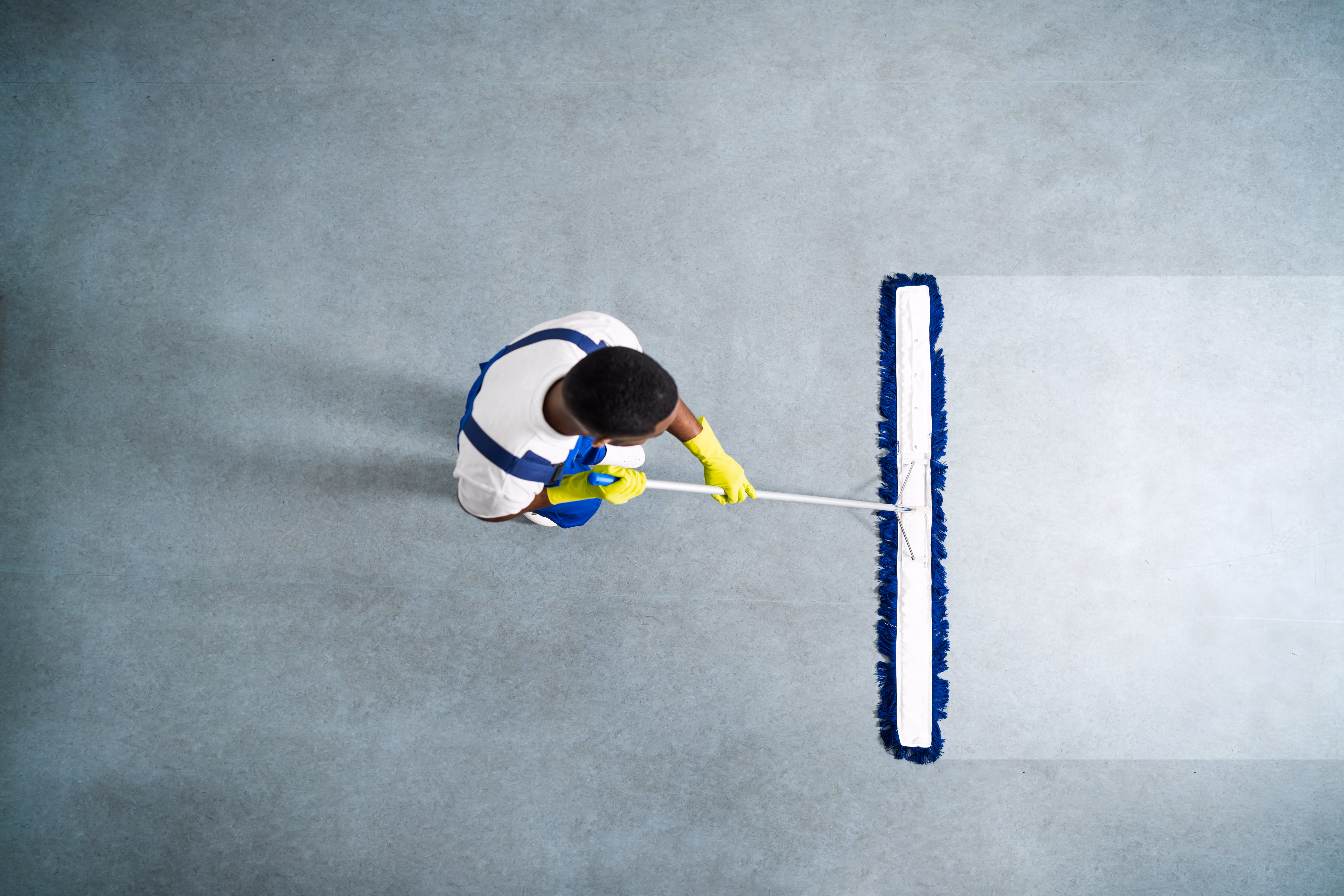 Top view of a person wearing yellow gloves and blue overalls cleaning a floor with a large mop.
