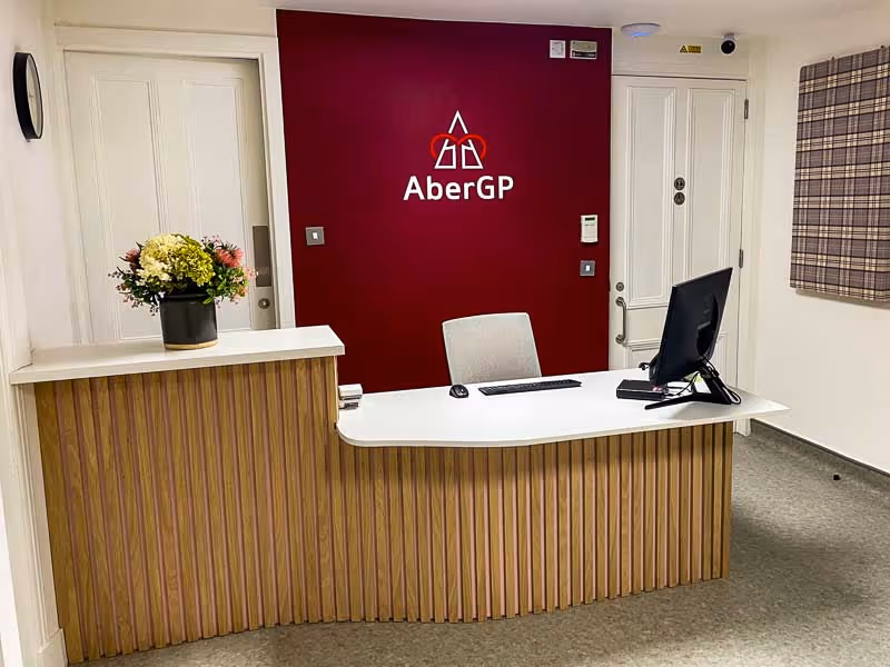 Reception desk with computer, chair, and flower arrangement in front of a burgundy wall with AberGP logo.