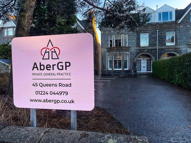 Pink sign for AberGP Private General Practice with address 45 Queens Road, phone number 01224 044979, and website www.abergp.co.uk, in front of a stone building and driveway.
