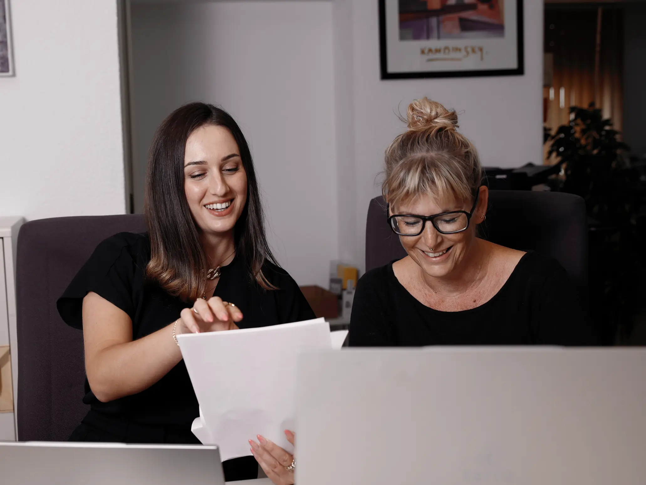 Zwei Frauen lächeln und betrachten gemeinsam Dokumente in einem Büro.