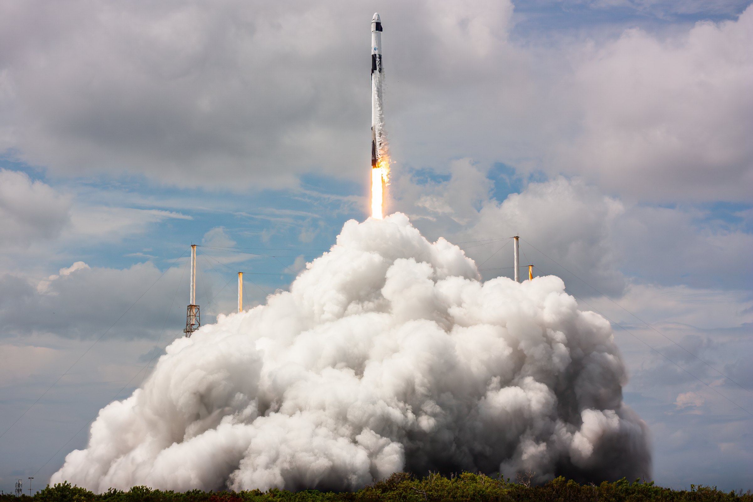 A rocket rises out of its own exhaust cloud into a broken cloud deck in daylight.