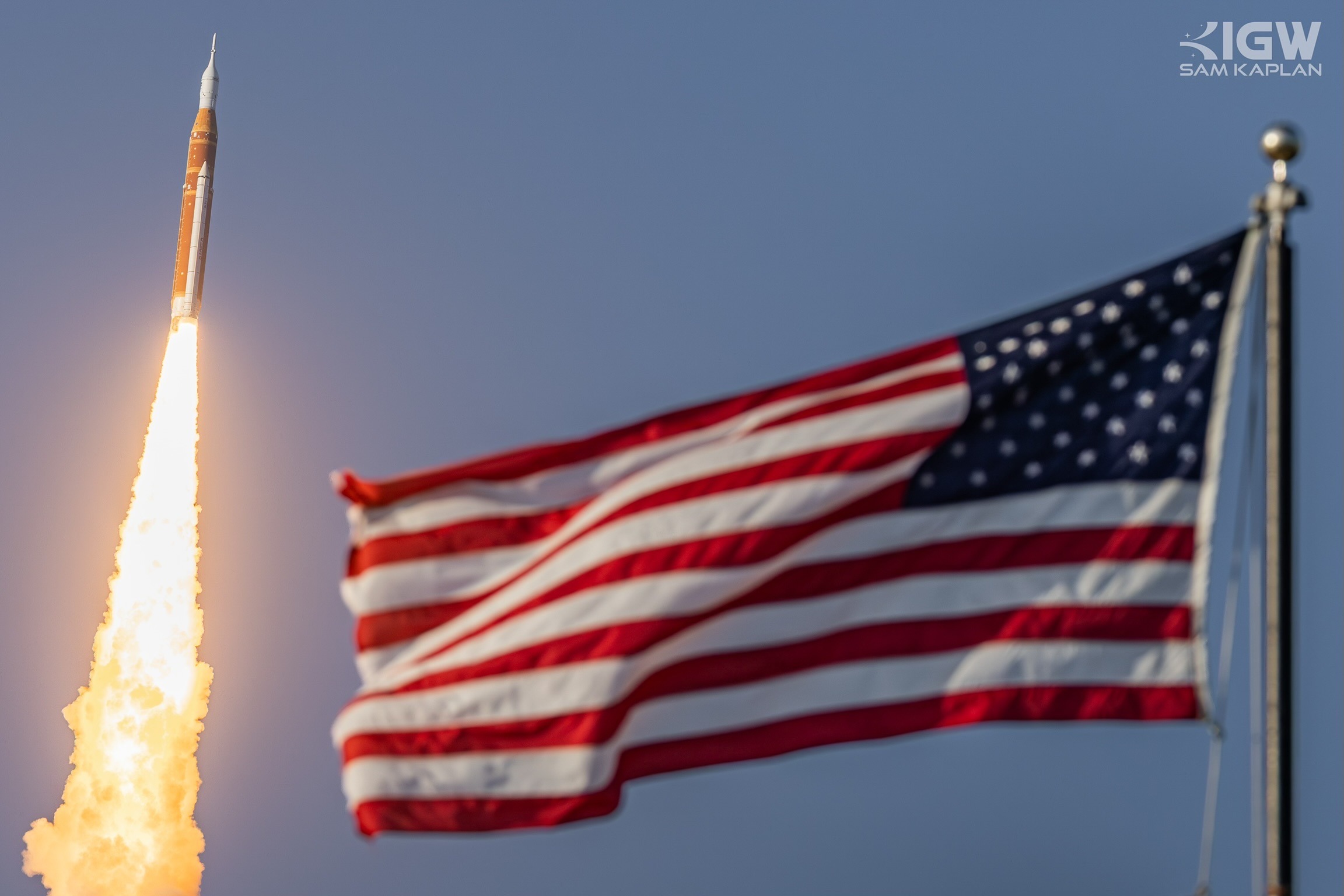 The Space Launch System climbs past a United States flag, its exhaust column bright against a clear sky.