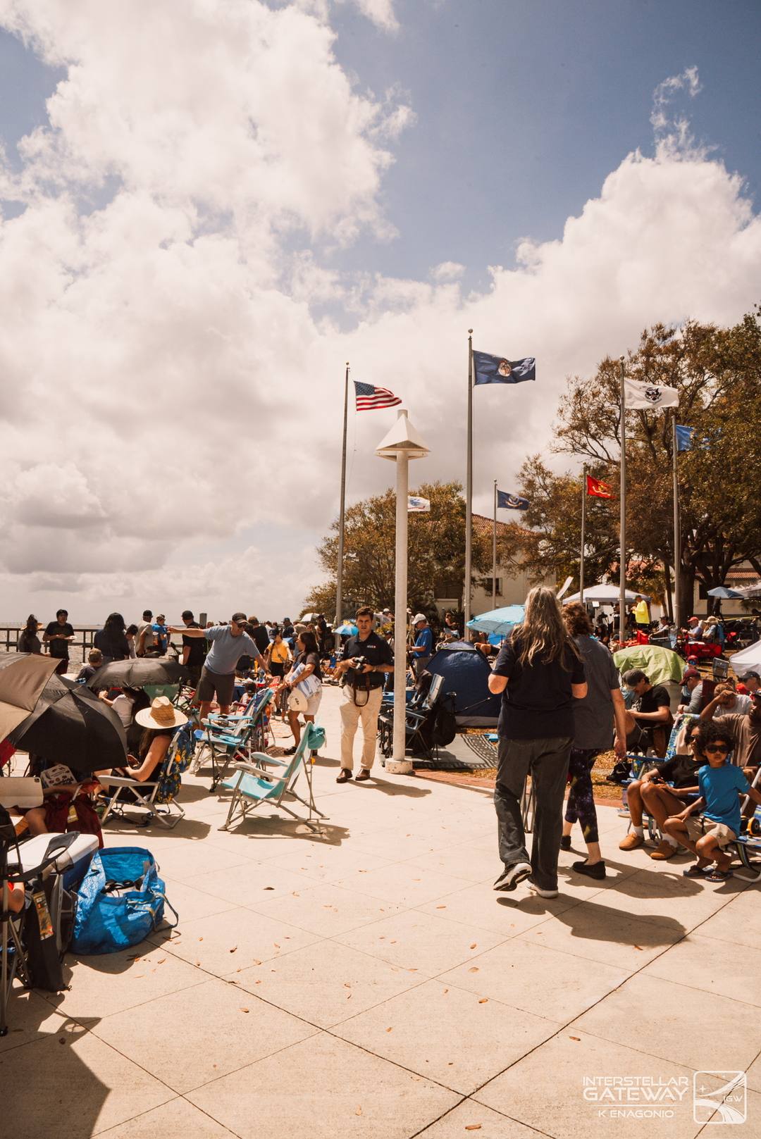 Spectators on a paved waterfront under a cloudy sky, flags flying above them.