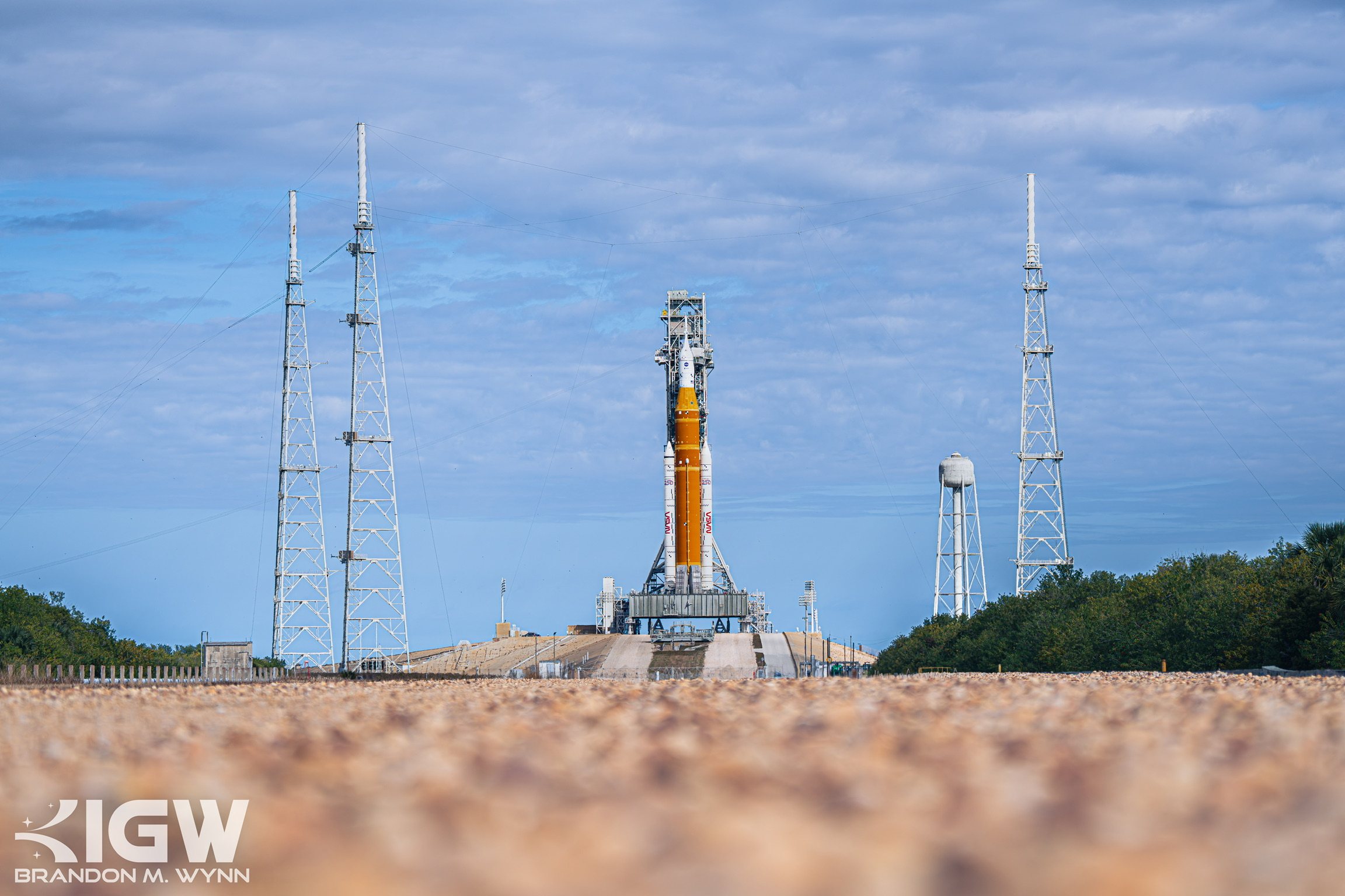 The Space Launch System on its mobile launcher seen from a distance across water and scrub, lightning towers either side.