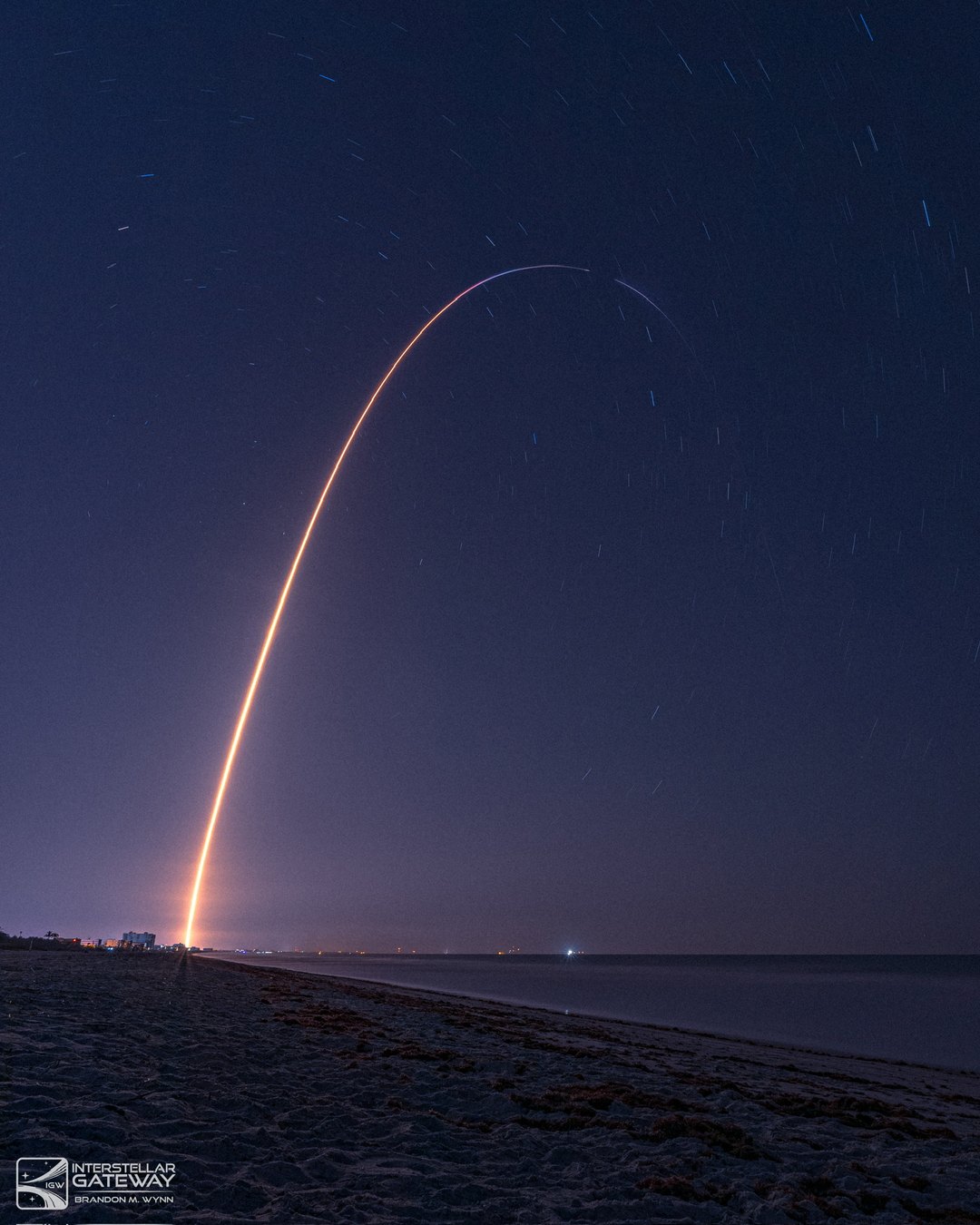 A launch arcs over the beach at night in a long exposure, the trail bending downrange.
