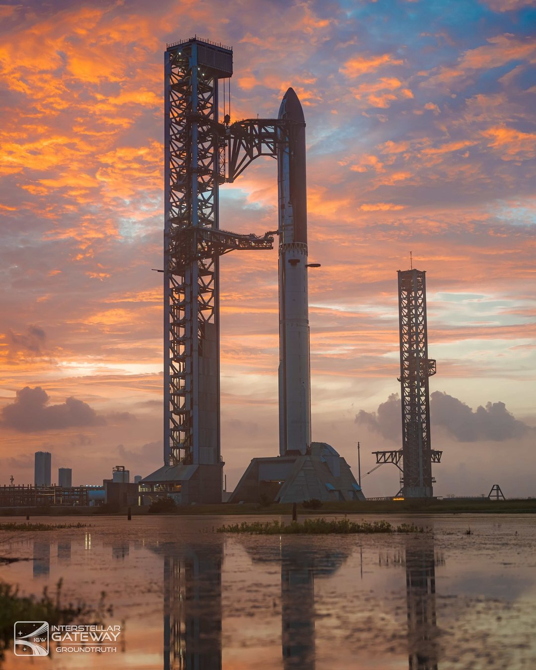 Starship and Super Heavy on the launch mount at Starbase at sunrise, reflected in standing water.