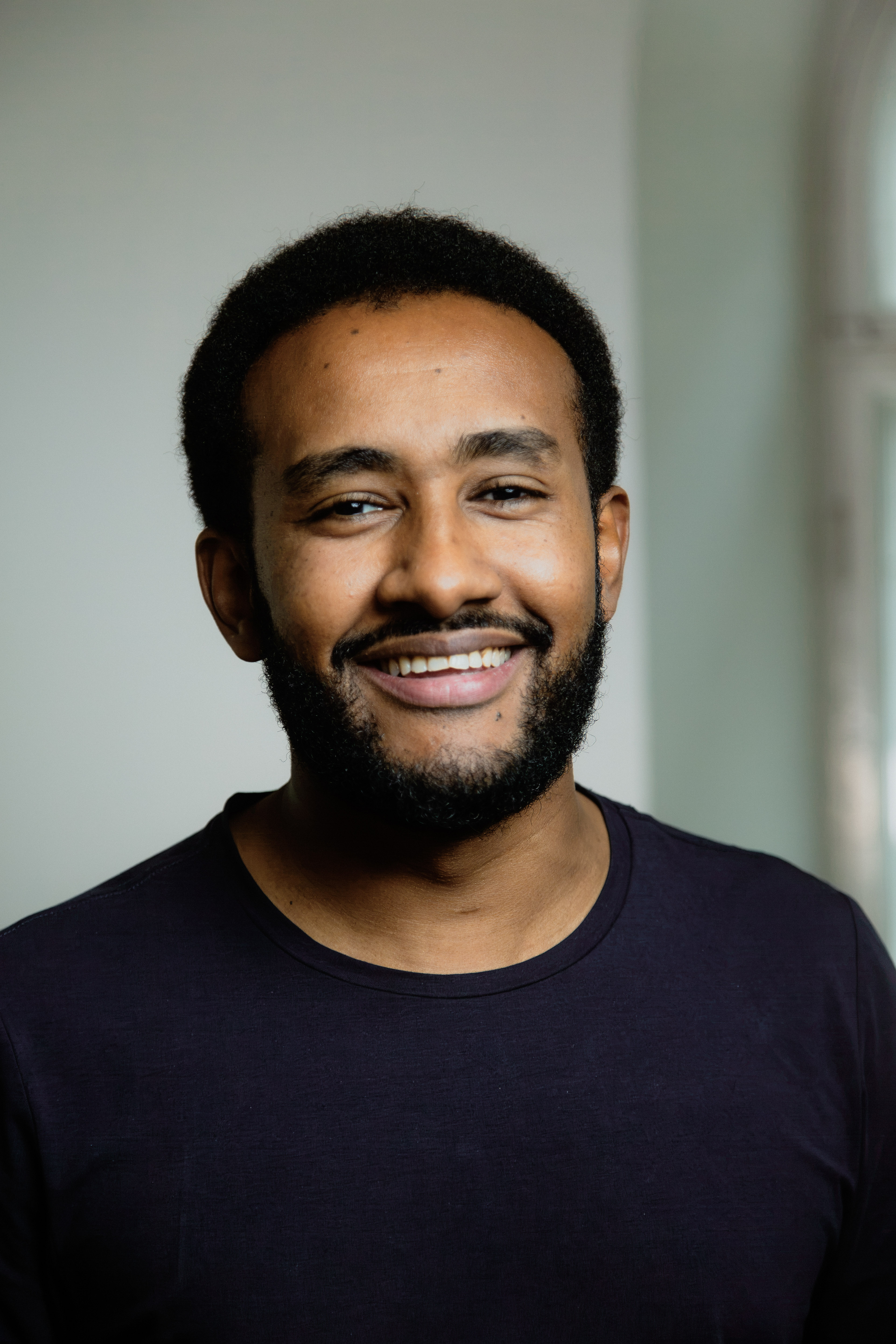 Smiling man with short curly hair and beard wearing a dark shirt against a light background.