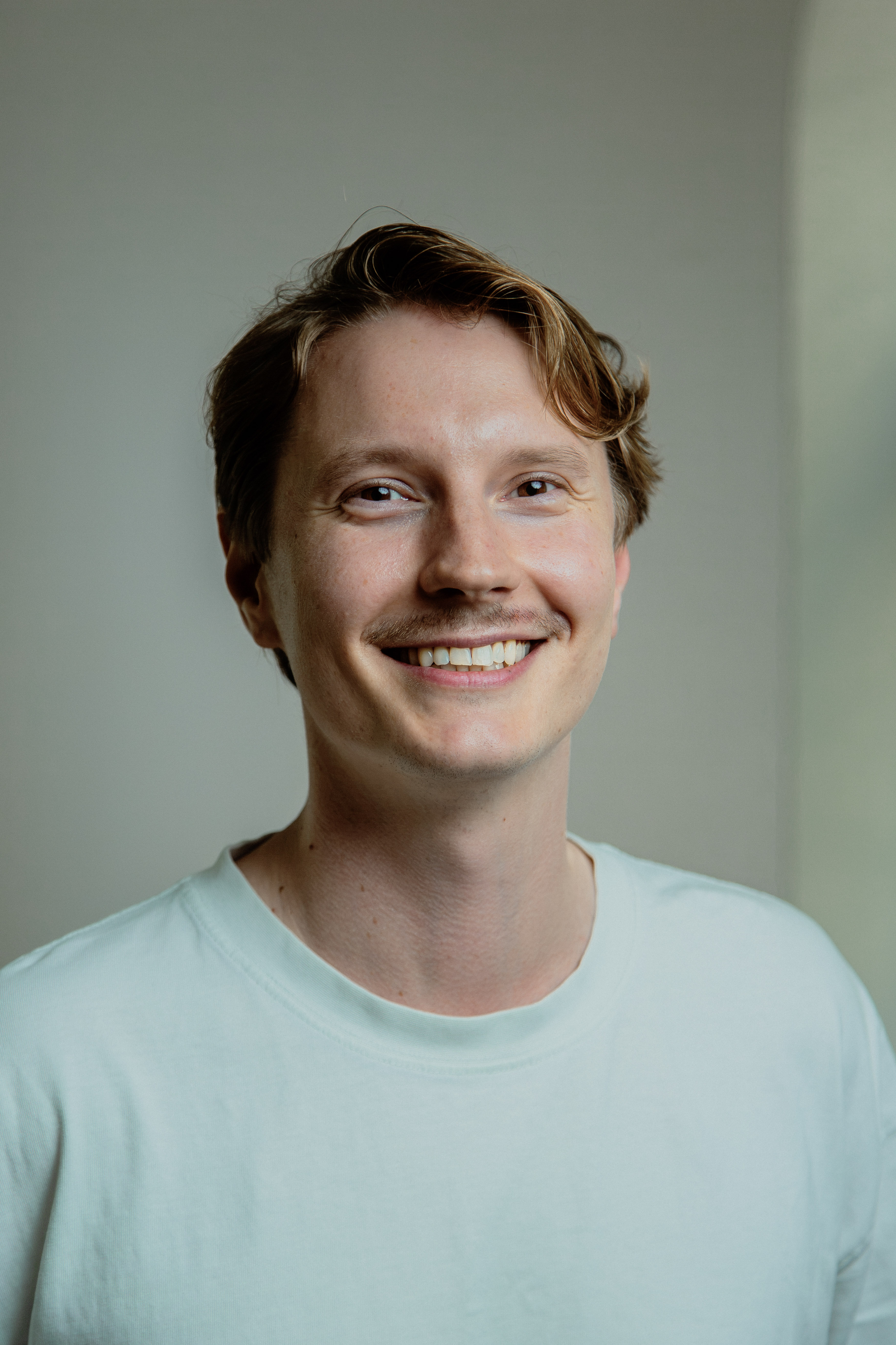 Smiling young man with light brown hair and mustache wearing a white shirt against plain background.