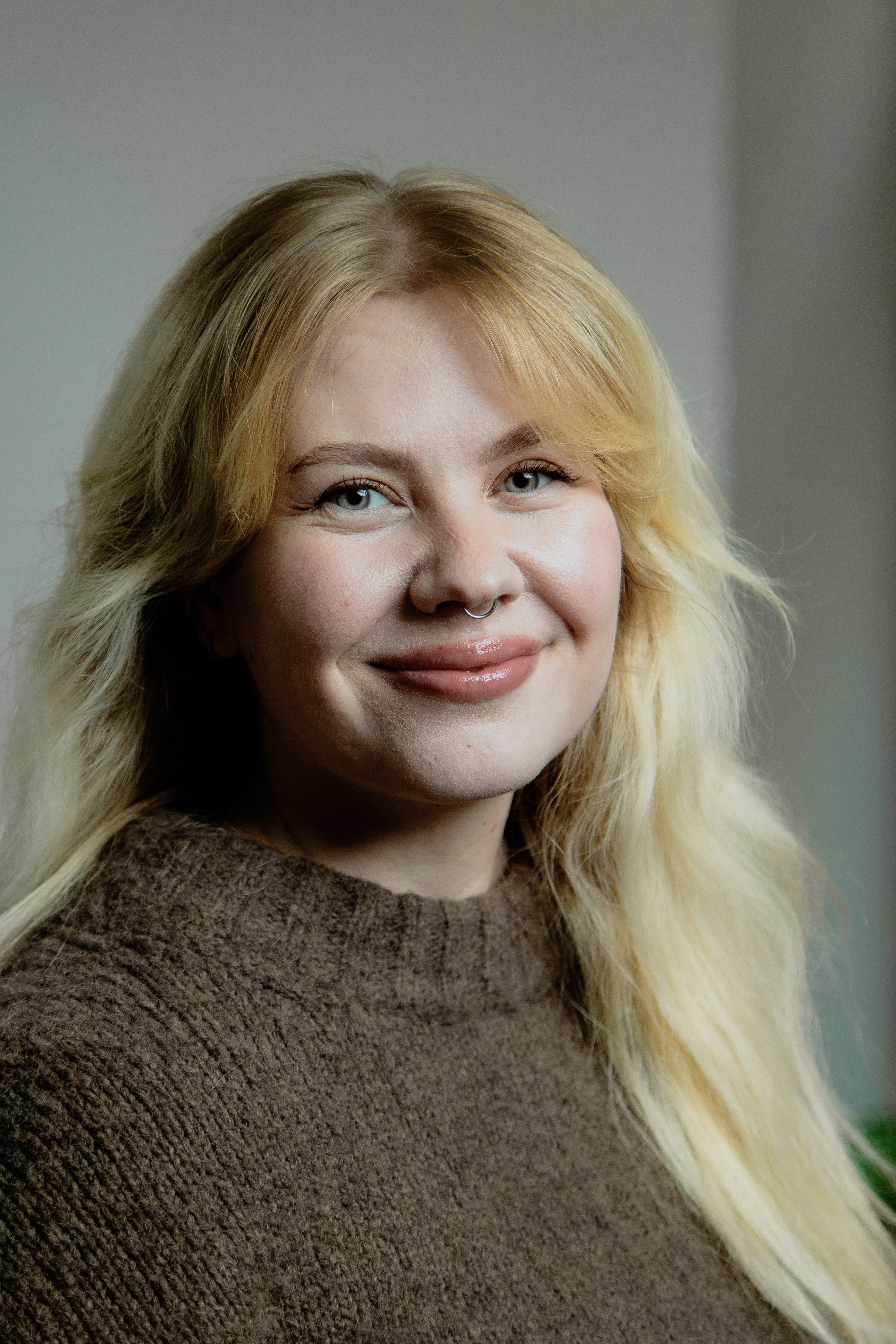 Smiling woman with long blonde hair, light blue eyes, and a nose ring wearing a brown knit sweater.