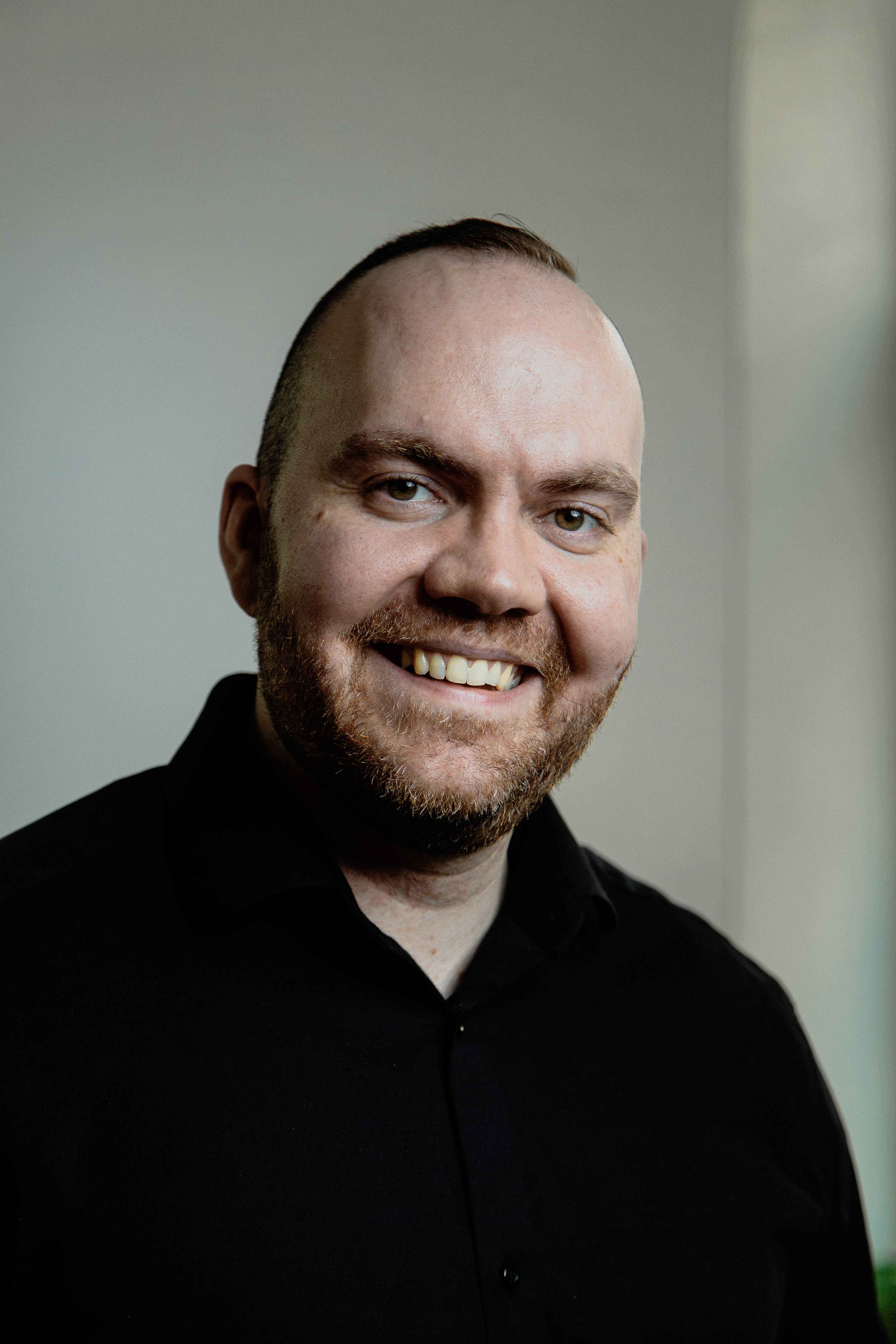 Smiling man with a beard and short hair wearing a black shirt against a plain background.
