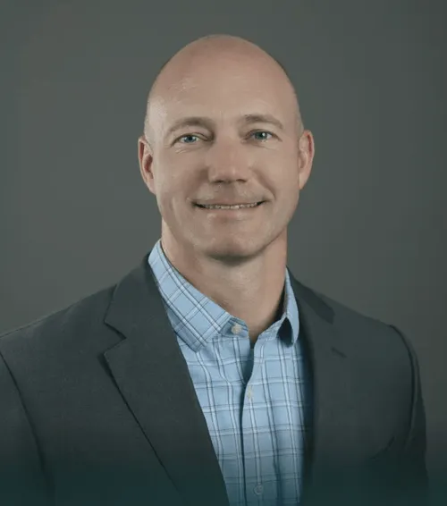 Smiling bald man wearing a dark blazer and blue checked shirt against a gray background.